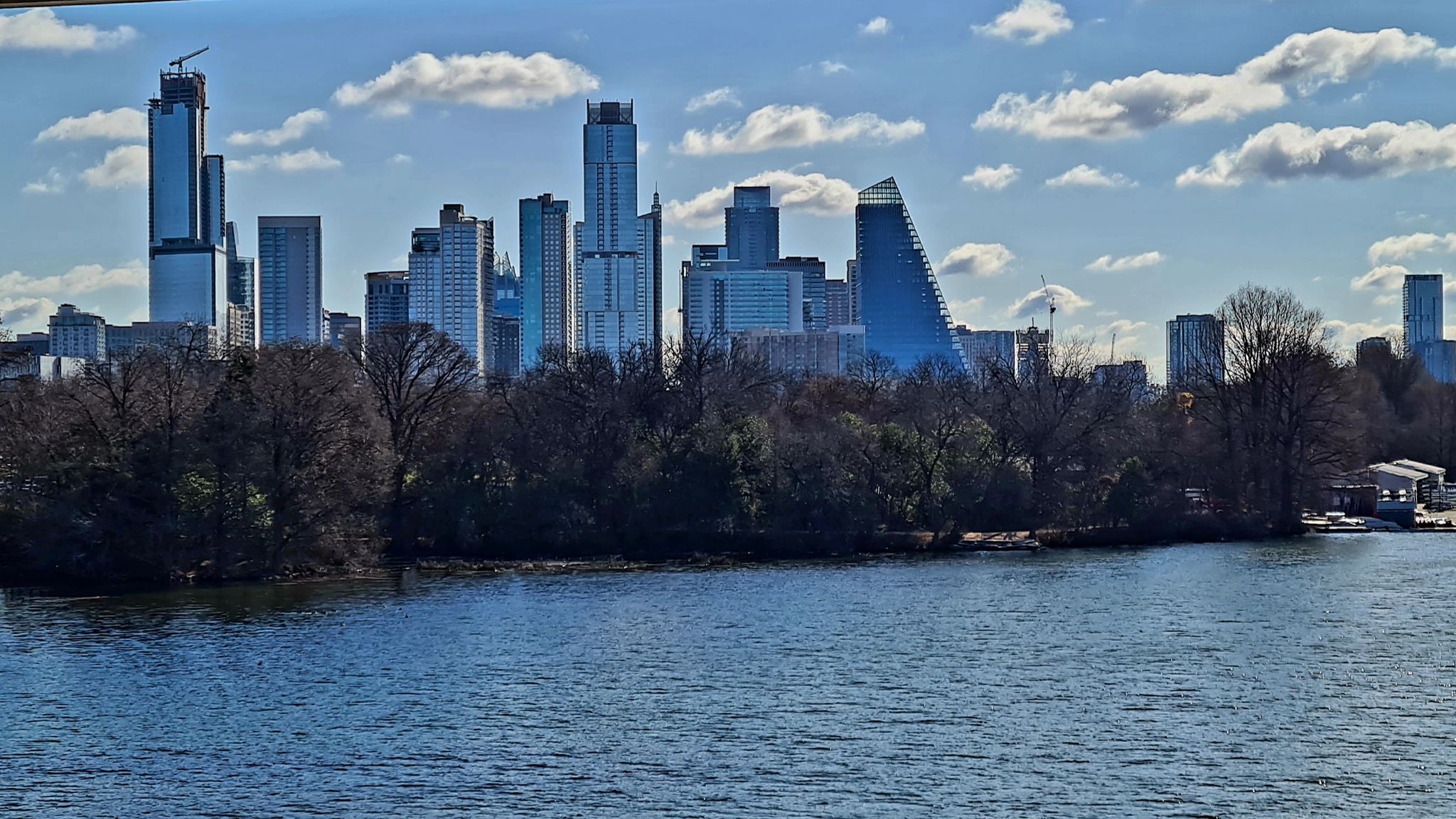 Modern city skyline behind a tree-lined riverbank under a cloudy sky. - Barton Springs Pool – Winding Path in Zilker Park loop from Zilker Eagle