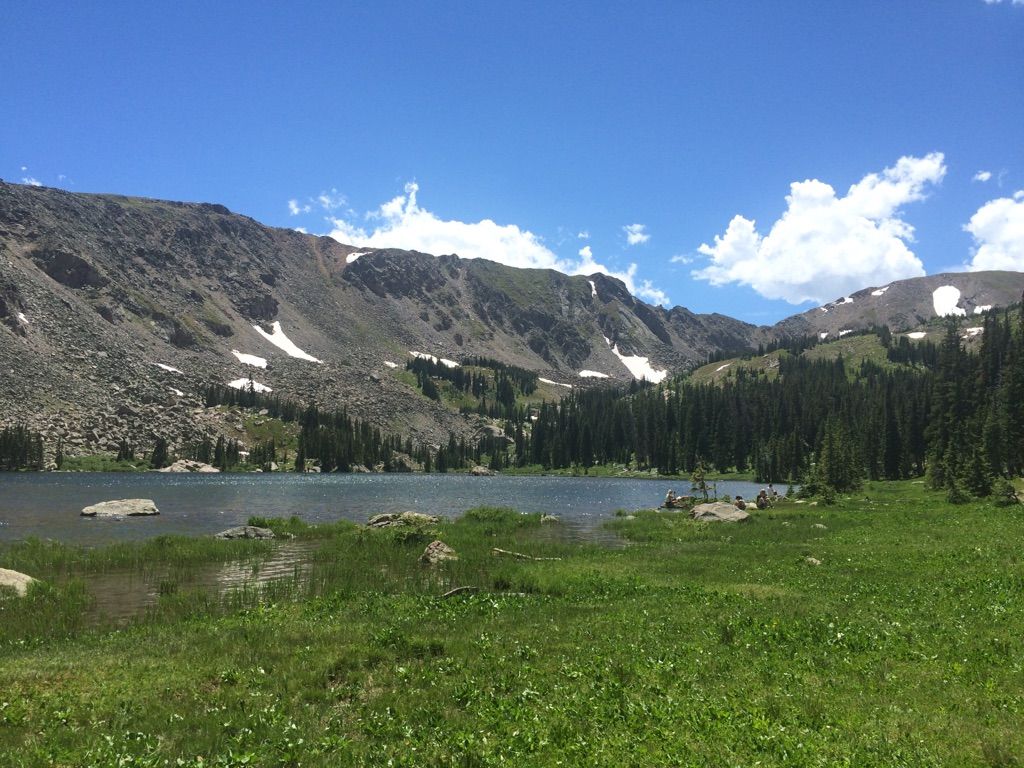A clear mountain lake with snow-capped peaks and green meadows on a sunny day. - Upper Diamond Lake Trail