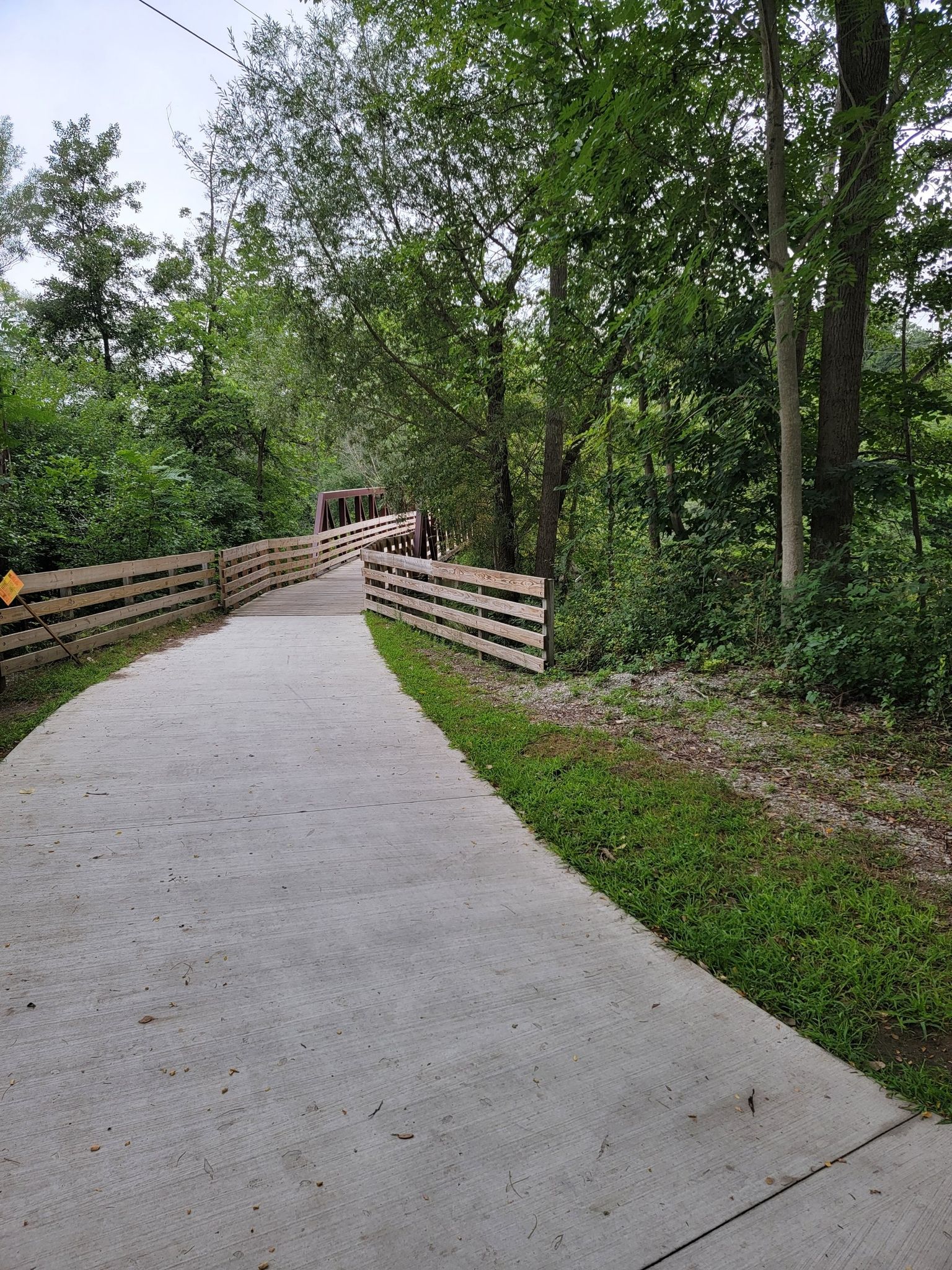 A paved path with wooden fences winds through a green forest towards a bridge. - Mary Beth Doyle Park Wetlands – Fuller Park loop from Arborland Center