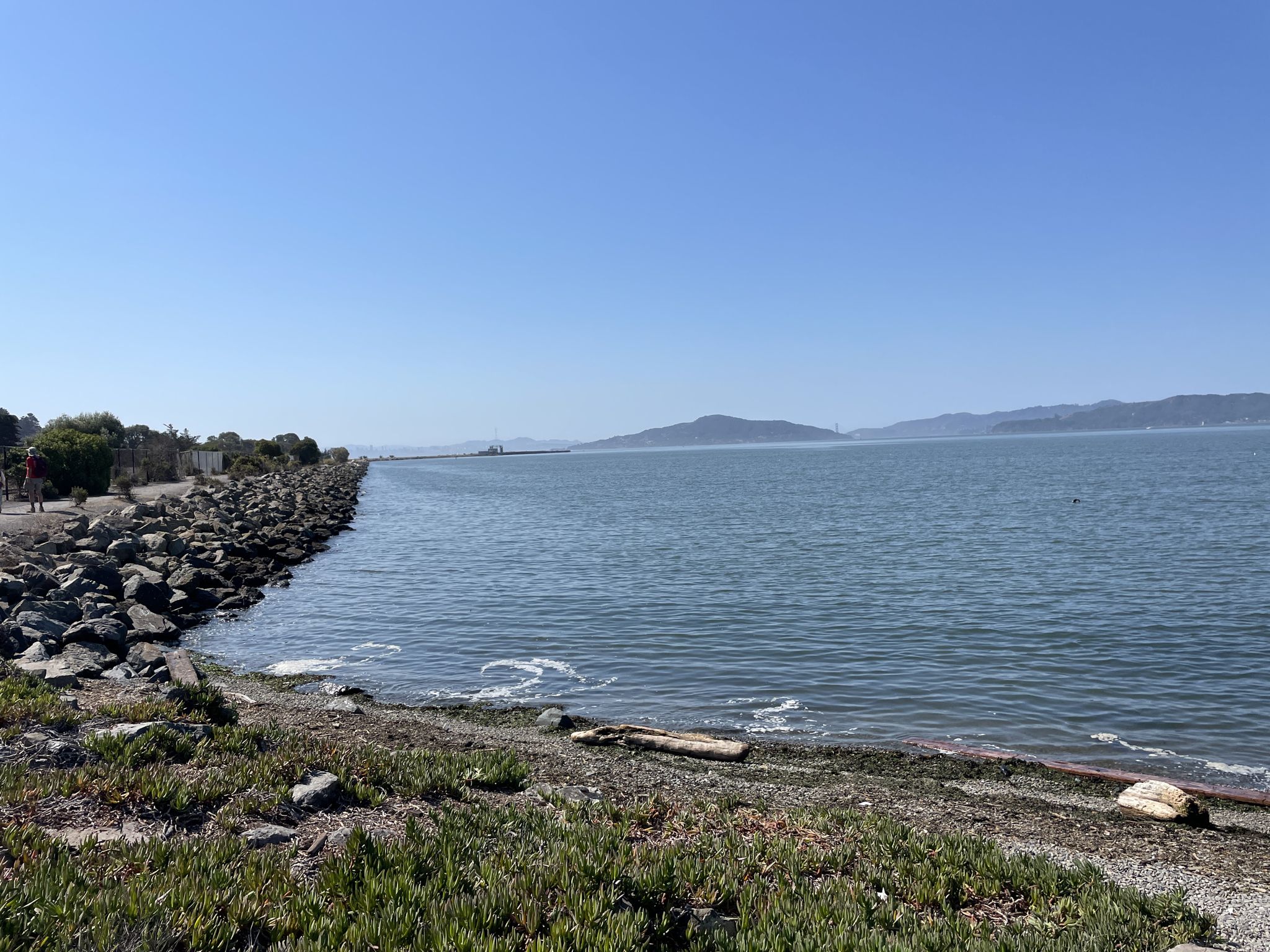 Rocky shoreline and calm bay water under a bright, clear blue sky. - Ferry Point Loop and Shipyard 3 Trail