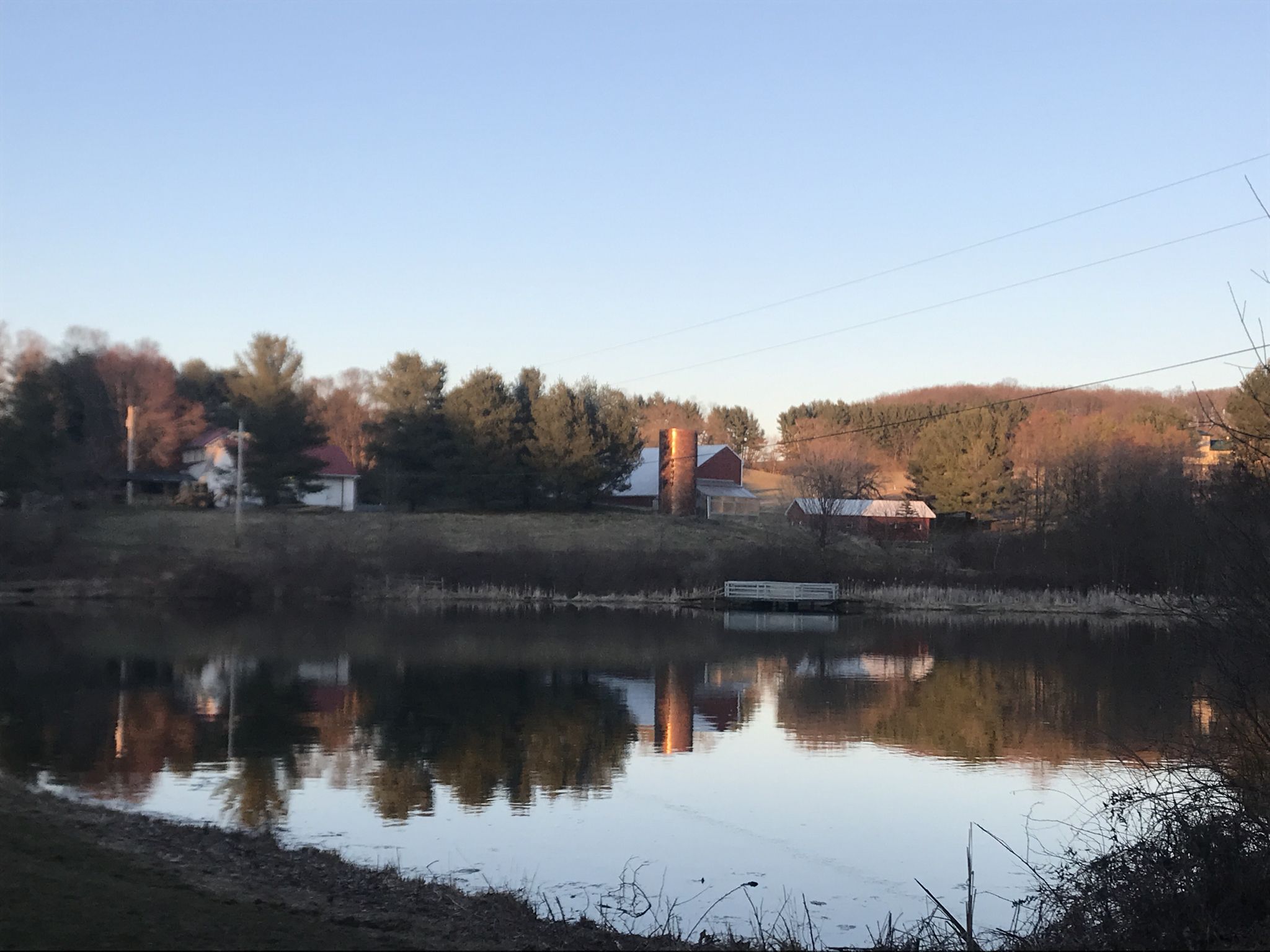 A serene pond reflects farm buildings and trees under a bright, clear sky. - Hashawha Loop Trail