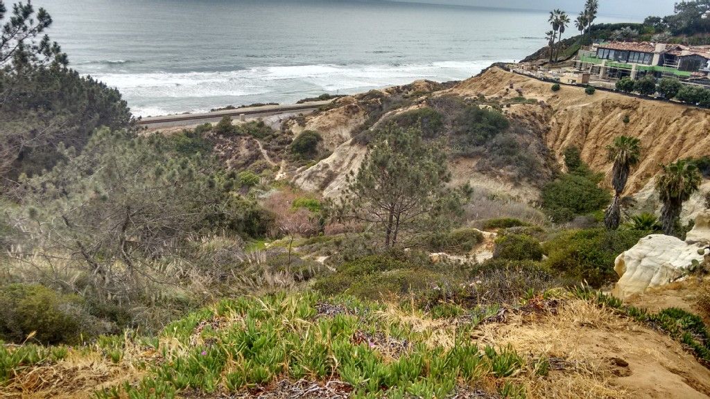 Overcast coastal view with ocean, sandy cliffs, green vegetation, and distant buildings. - California Mission Trail - stage 01 Mission San Diego de Alcalá (1769) to Mission San Luis Rey (1798)