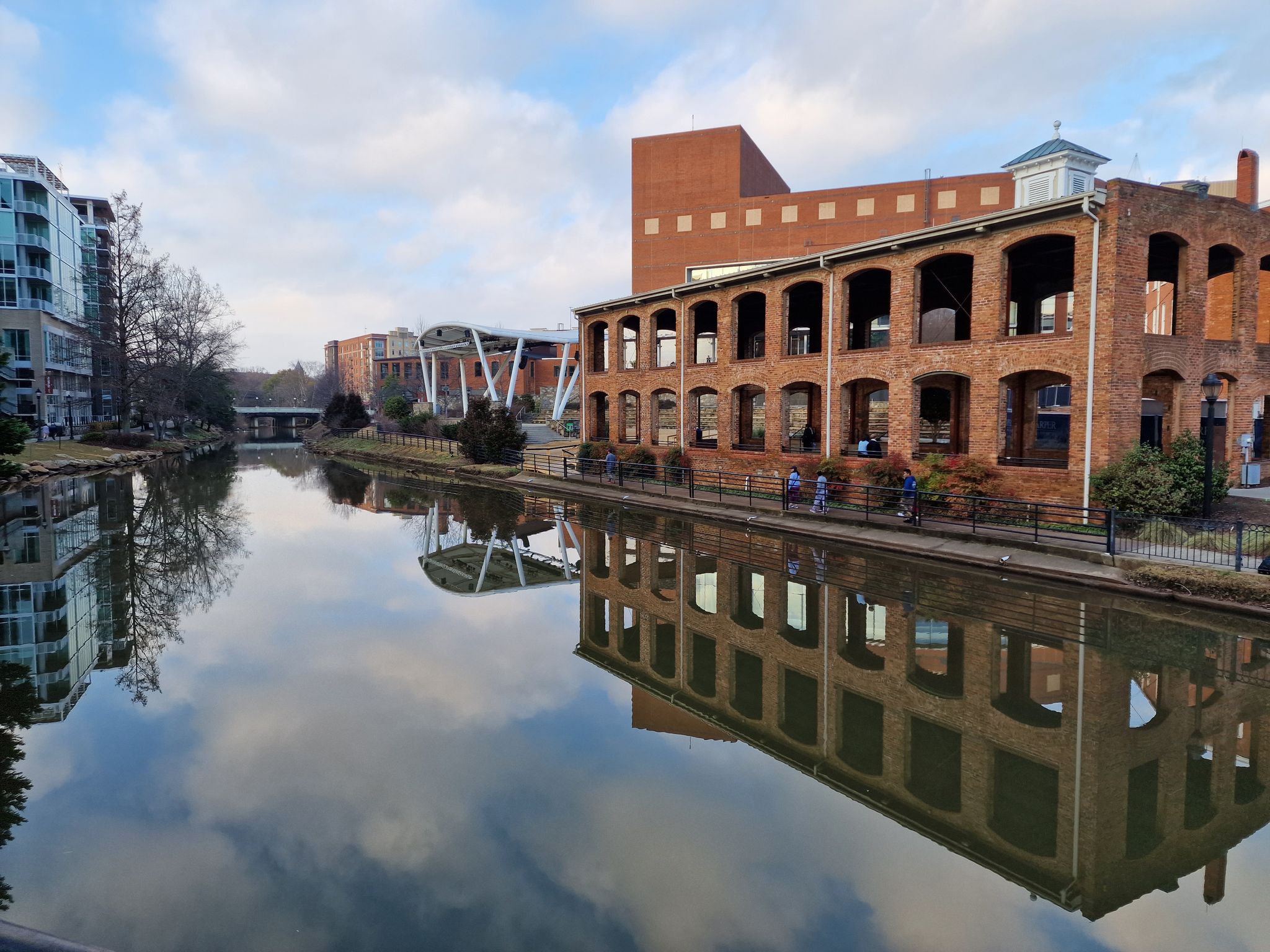 Calm river reflecting buildings and sky, with a brick building on the right. - Swamp Rabbit Trail and West End Loop