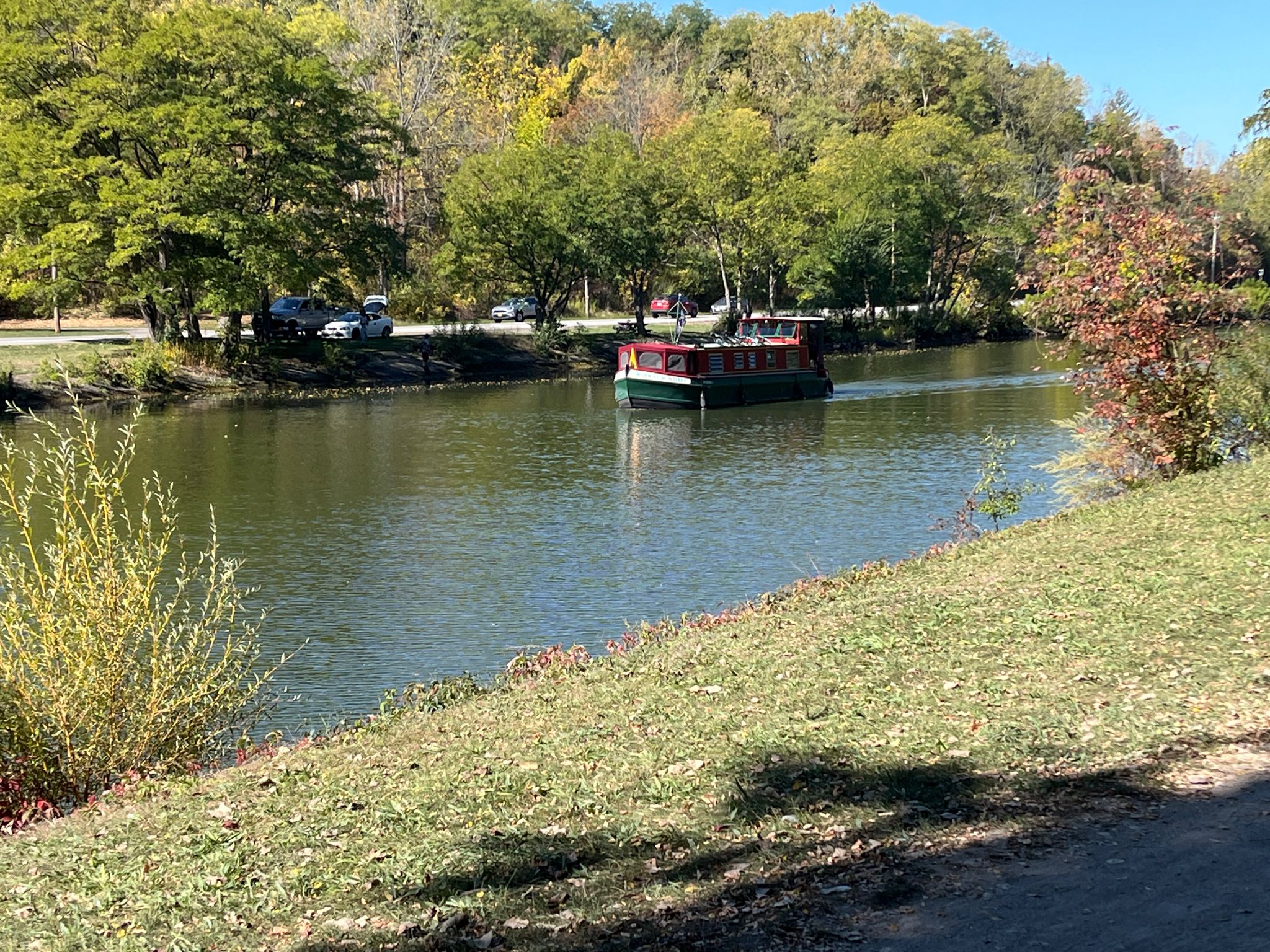 A boat moves on a river with trees showing autumn colors, sunny. - Pittsford loop from Bushnell's Basin