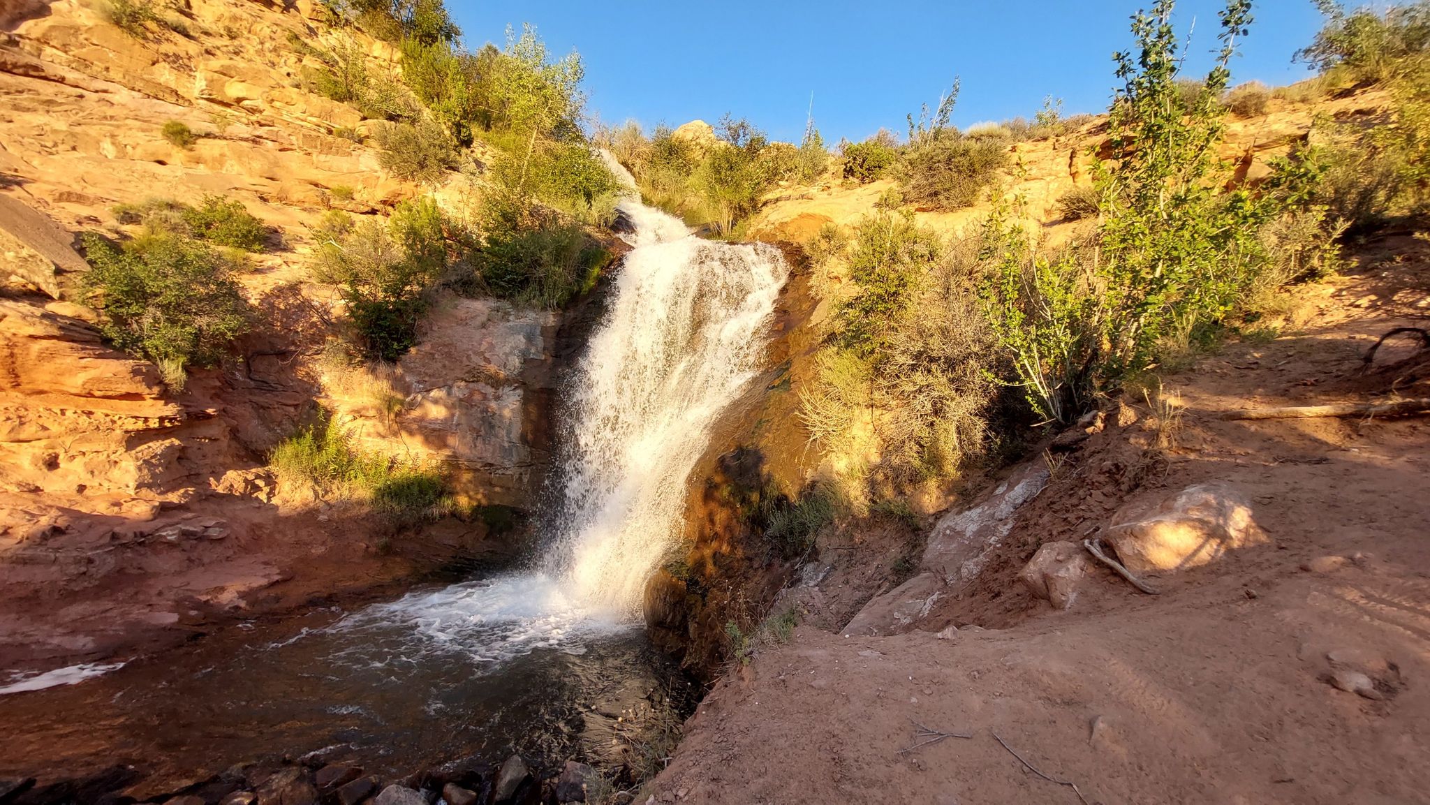 A waterfall cascades down reddish cliffs with sparse green vegetation under bright sunlight. - Ken's Lake and Faux Falls Trail