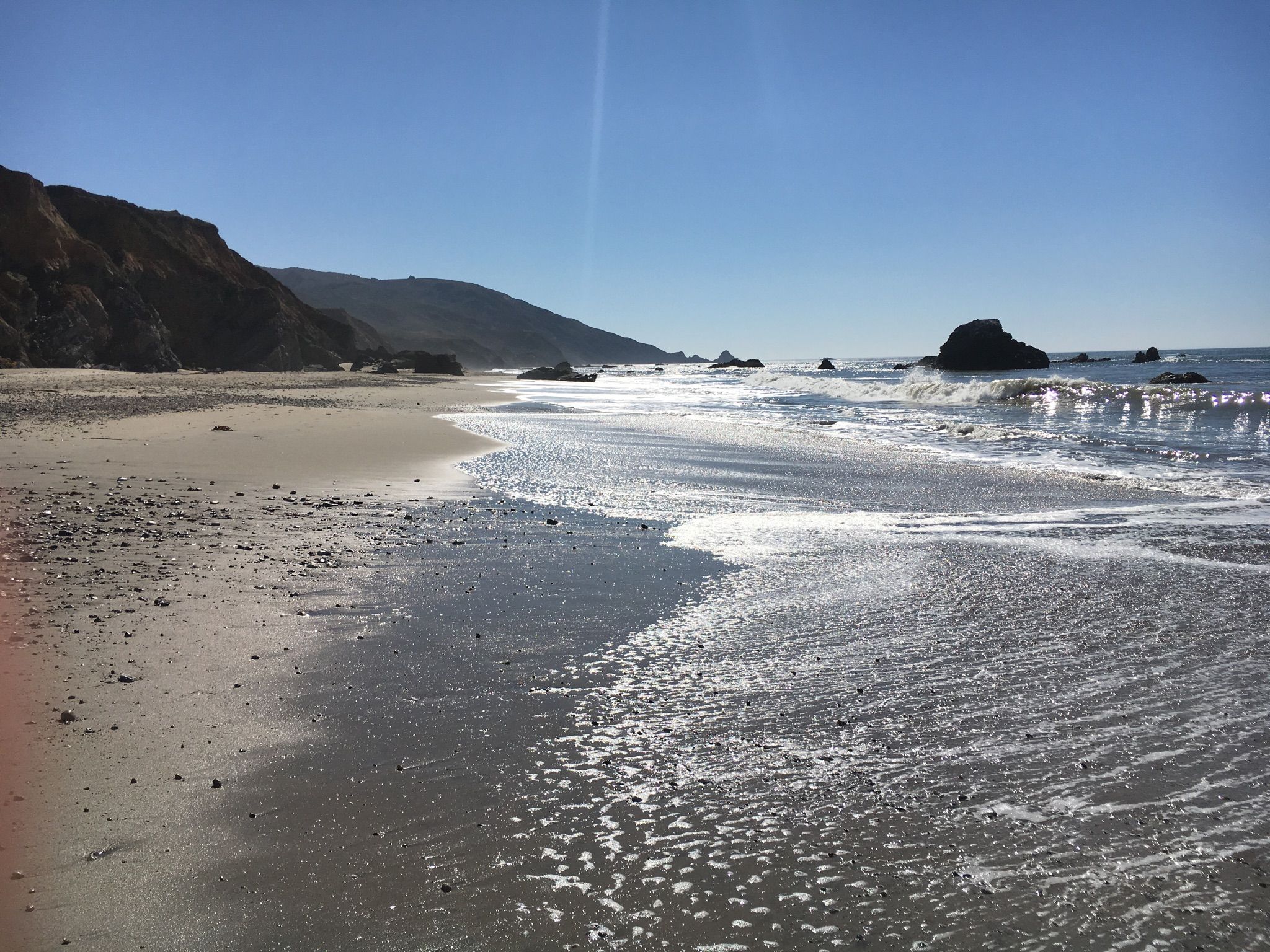 Sunny beach with foamy waves, rocky shore, and distant coastal hills. - Bobcat Trail – River Trail Loop