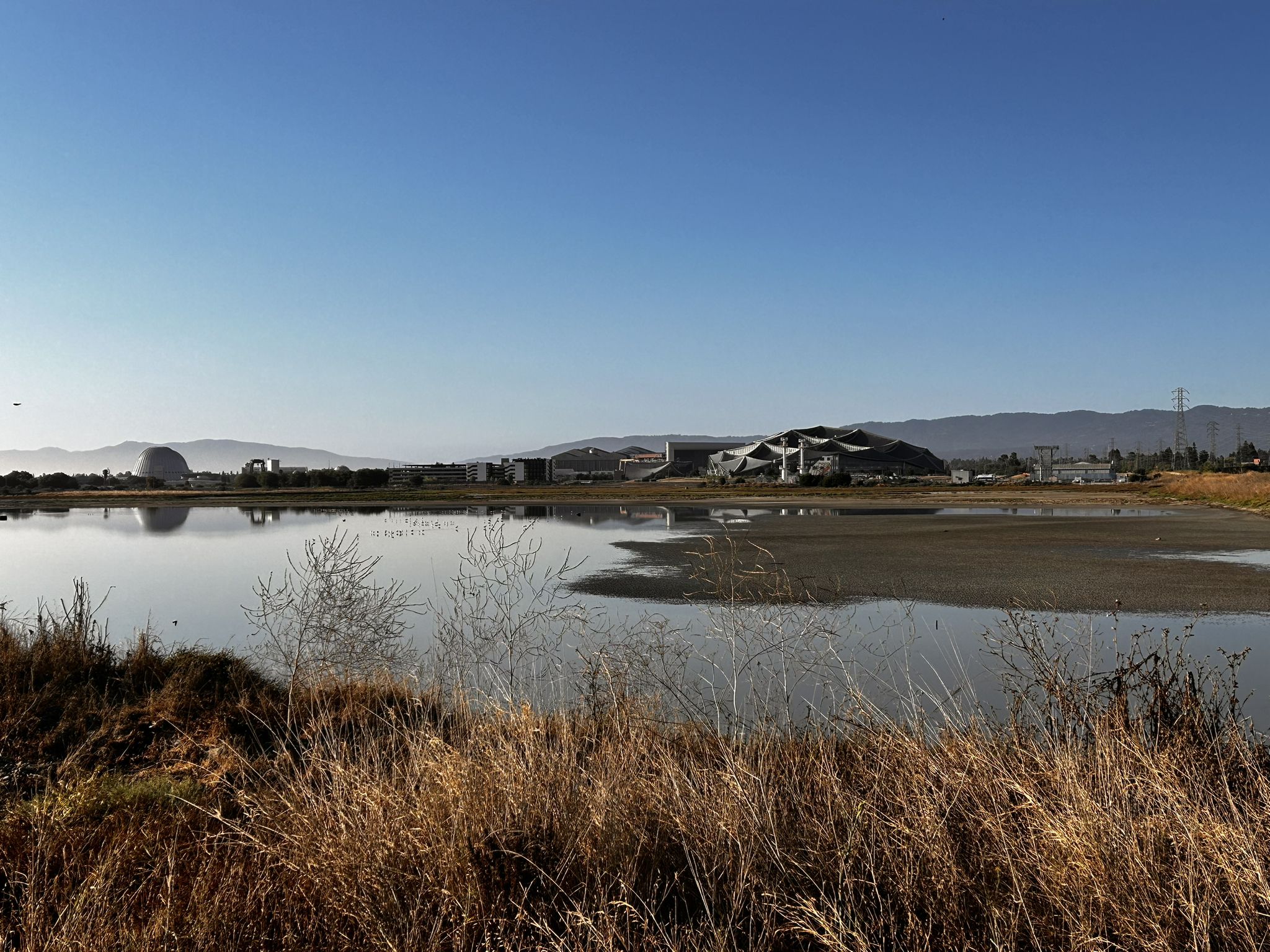 Still water reflects distant modern buildings and mountains under a clear blue sky. - San Francisco Bay Trail