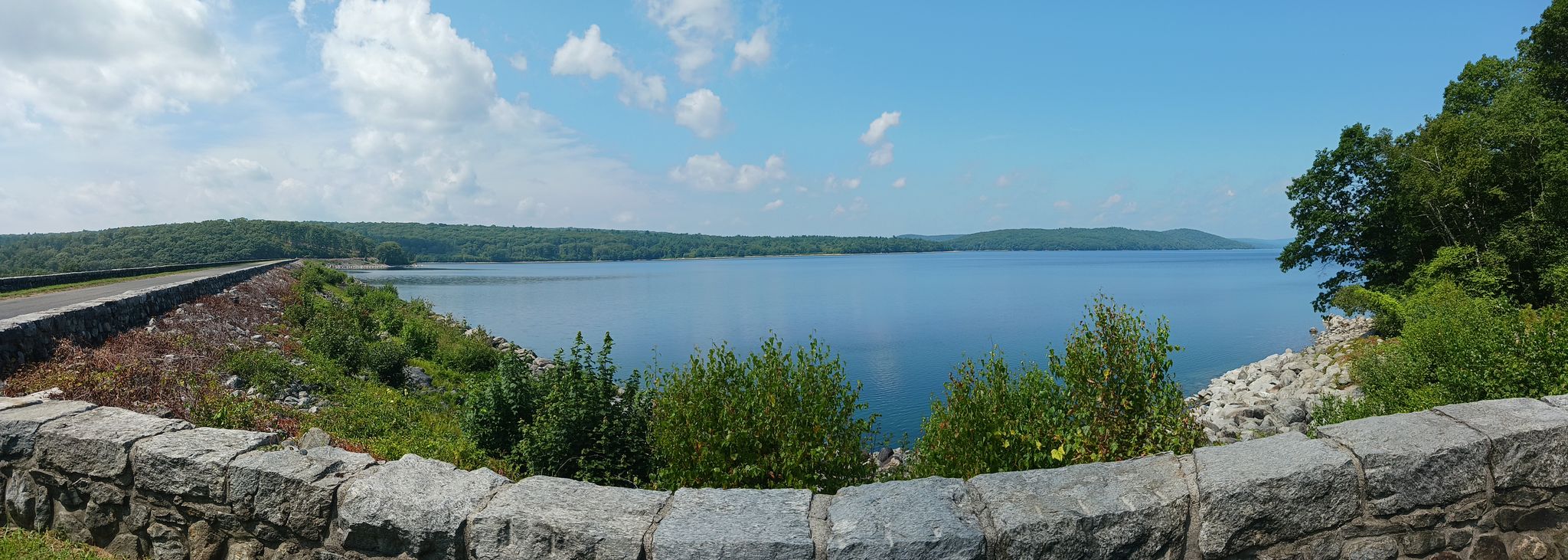 Panoramic view of a calm lake from behind a stone wall on a bright day. - Goodnough Dike Loop