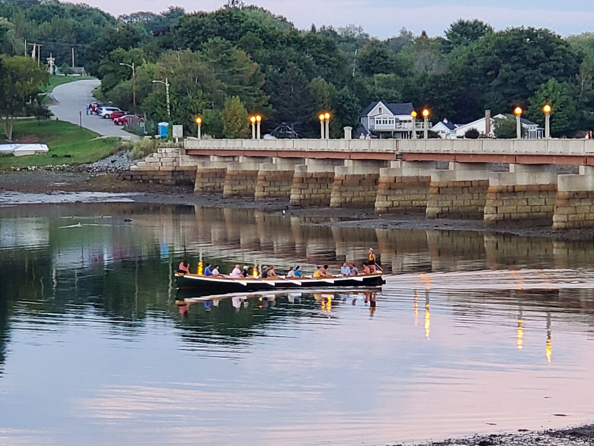 Long boat with many people rowing on water near a lit bridge at dusk. - Start of Hawthorne Hike – Belfast Rail Trail loop from Belfast