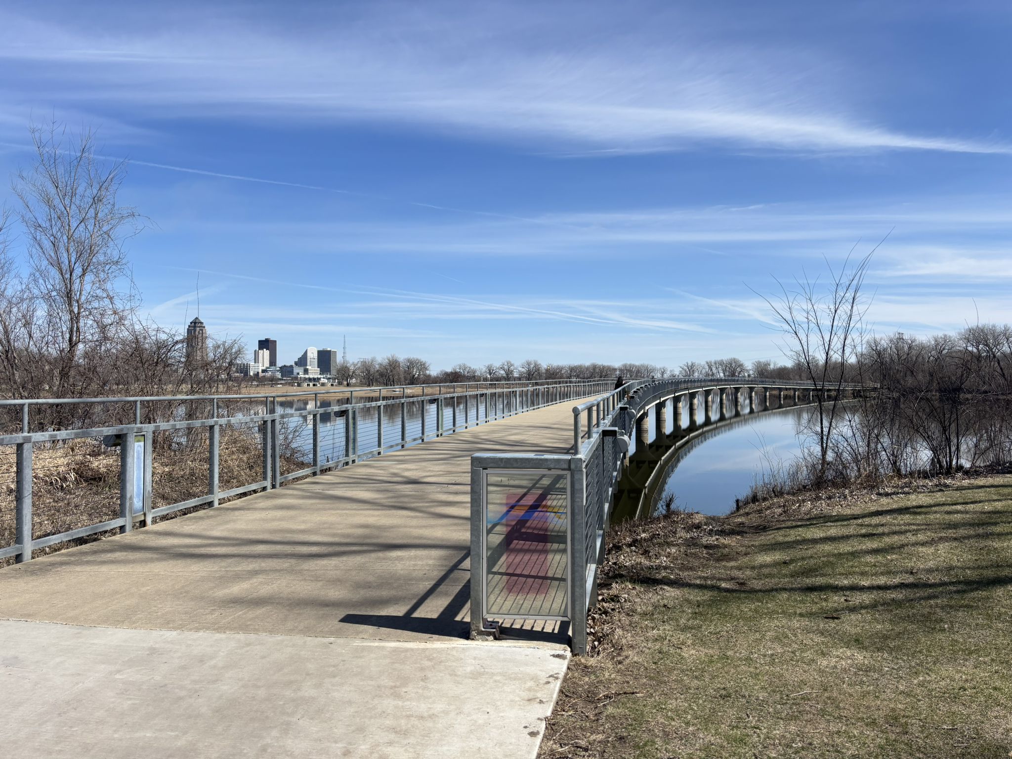 A long modern bridge spans calm water, with a distant city skyline under blue sky. - Gray's Lake beach – Gray's Lake Park loop from Des Moines