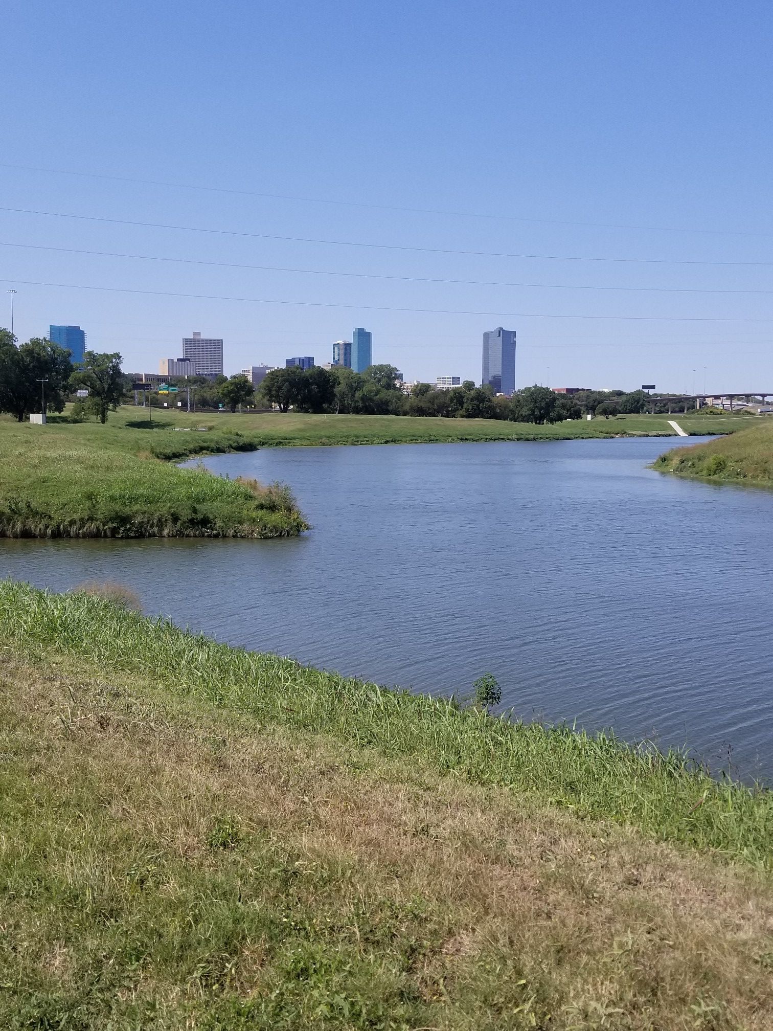 A winding river with grassy banks under a clear sky, city skyline in distance. - Trinity River – Henderson Street Bridge loop from Texas Wesleyan University