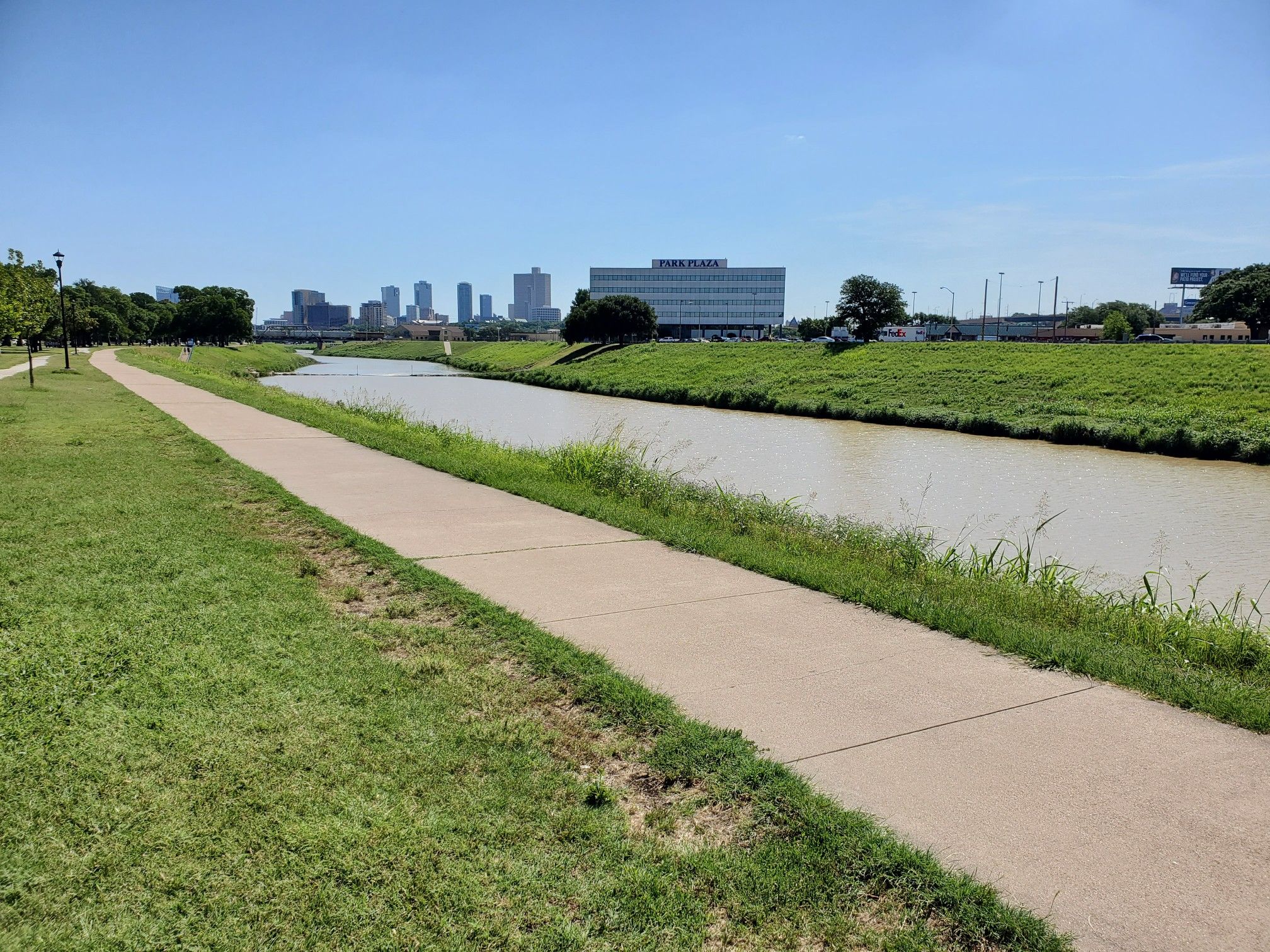 A paved path runs alongside a river with distant city buildings. - Trinity Park – Henderson Street Bridge loop from Texas Christian University