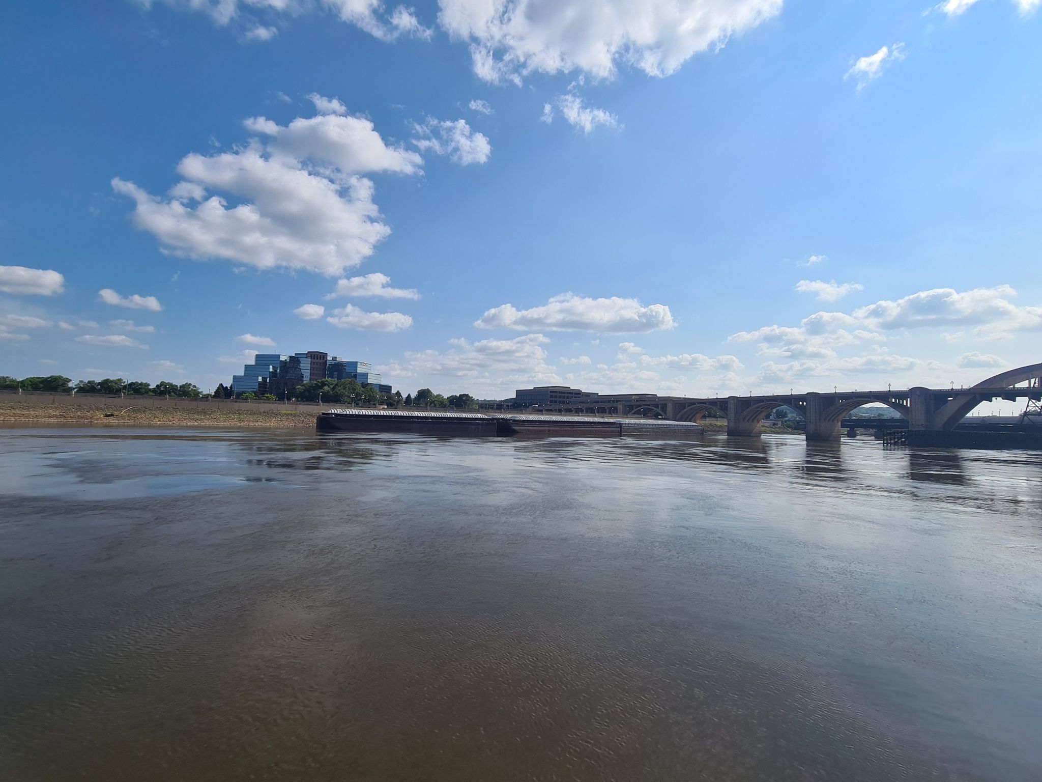 Wide river with a barge and distant bridge under a bright blue sky. - Samuel H. Morgan Regional Trail