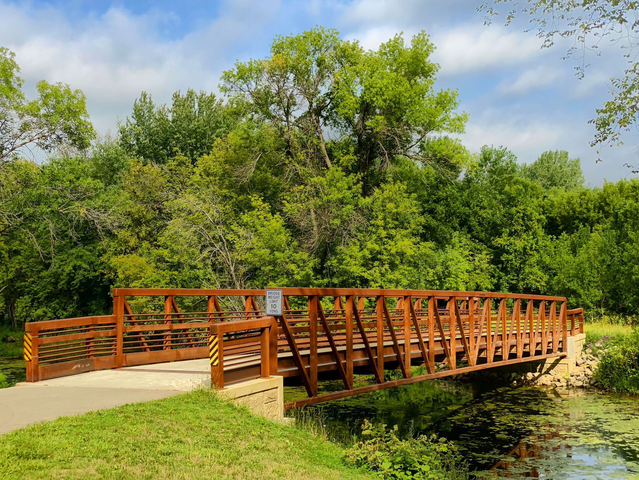 A wooden bridge spans a calm stream surrounded by lush green trees on a sunny day. - Minnesota Valley State Trail