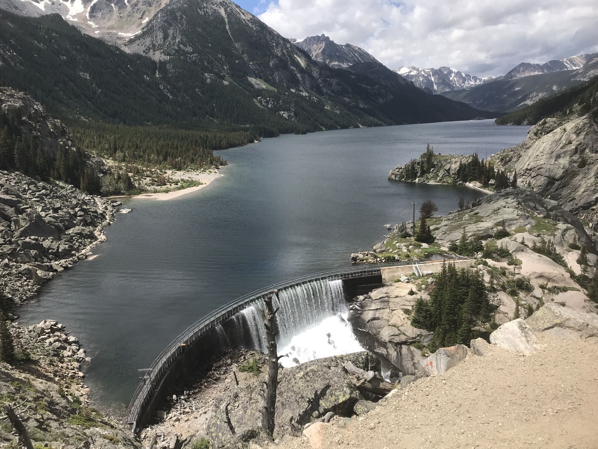 Expansive lake with a dam and waterfall, surrounded by rugged mountains under cloudy sky. - Mystic Lake via West Rosebud Trail