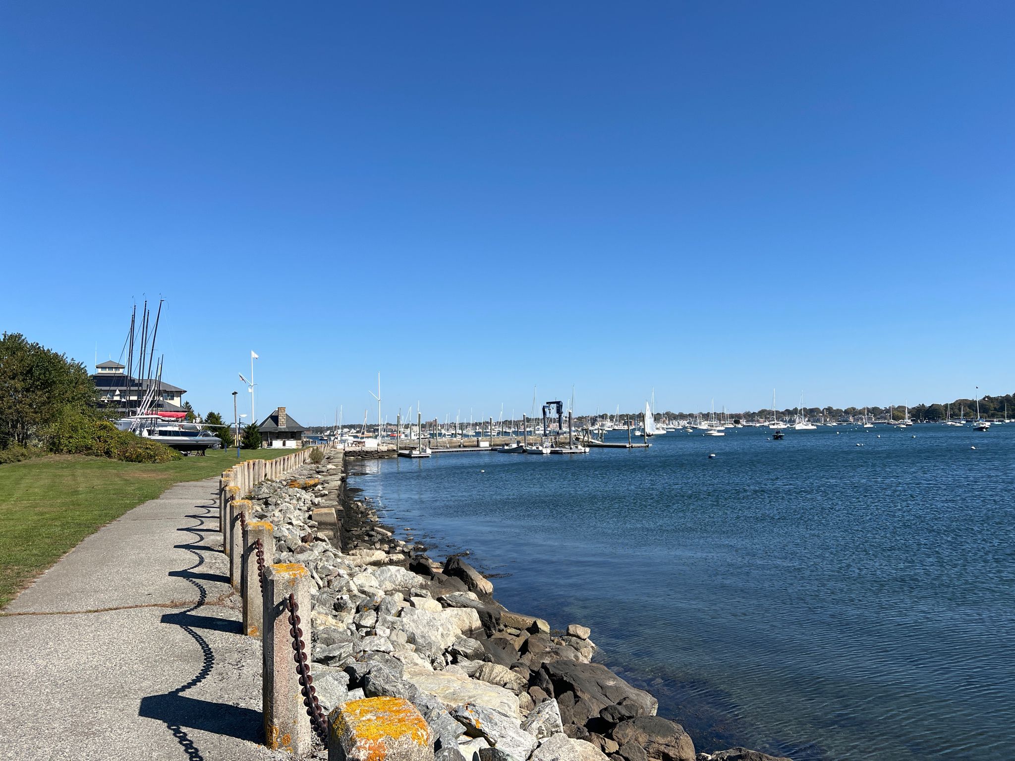 A paved path runs along a rocky shore with many boats in a sunny harbor. - Portuguese Discovery Monument – Fort Adams State Park loop from Fort Adams State Park