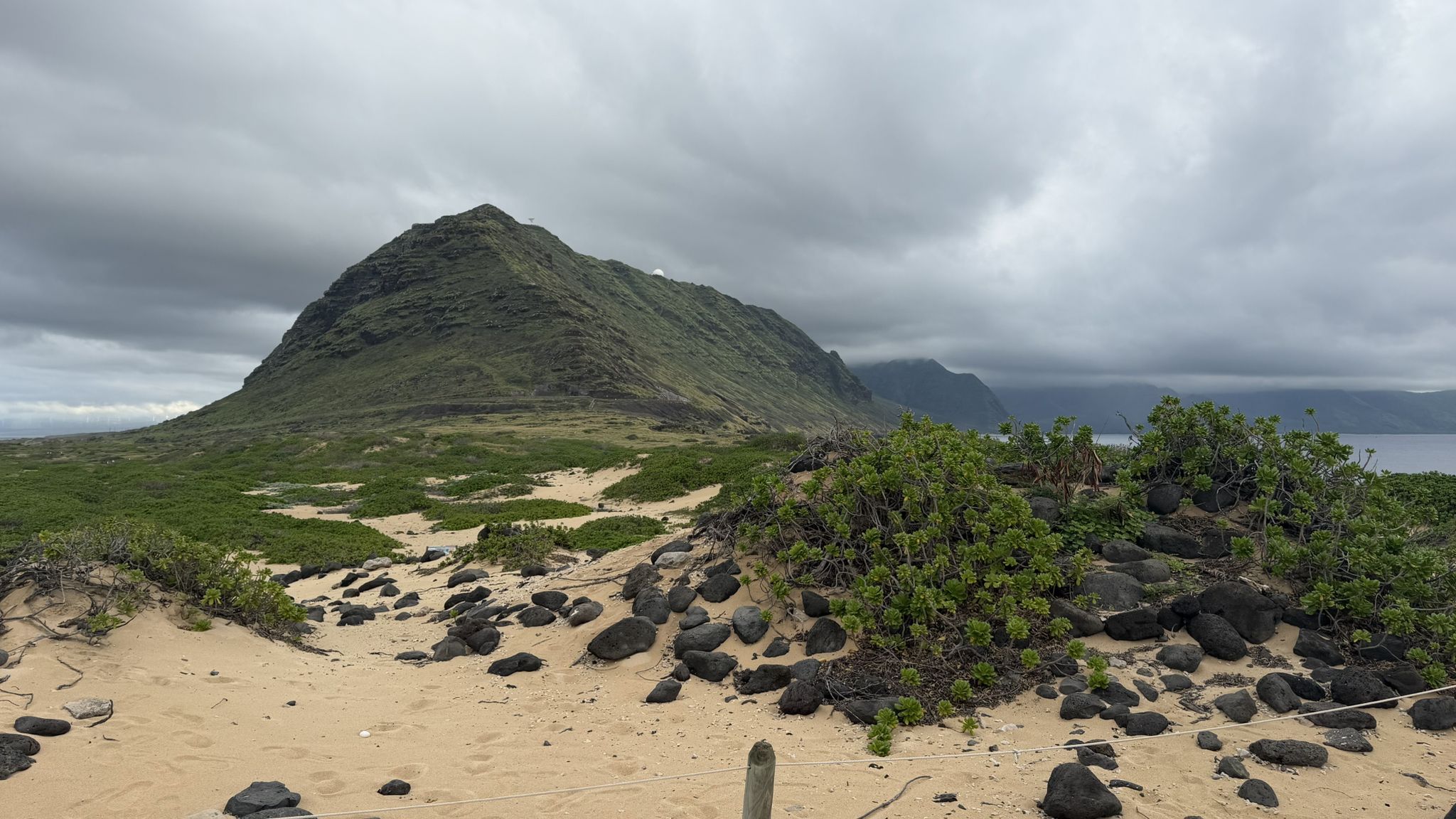 Large green mountain overlooking sandy terrain with dark rocks under cloudy sky. - Sea arch at Kaʻena Point – Westernmost Point of Oʻahu loop from Kaena Point Satellite Tracking Station