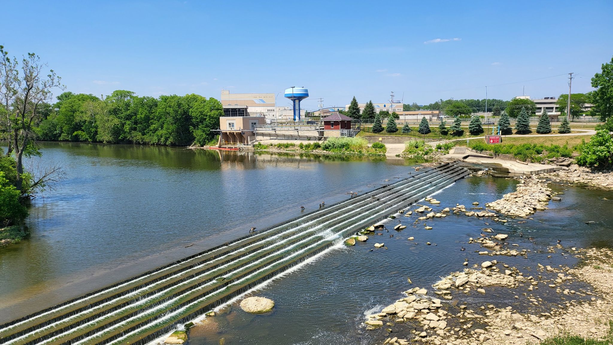 River with a dam, cascading water, buildings, and trees under a clear sky. - Scioto and Olentangy Trail Loop