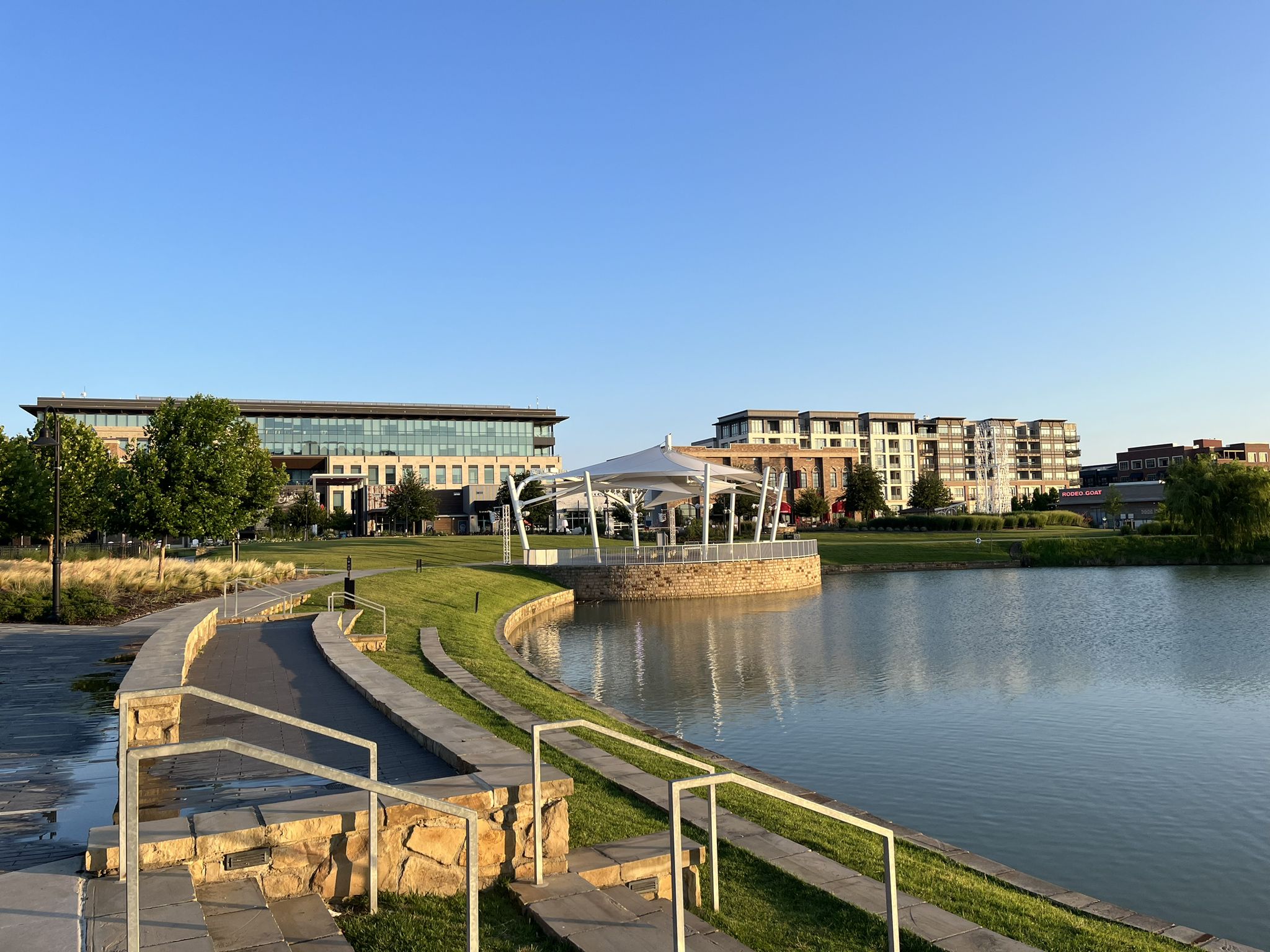 Modern buildings overlook a calm lake with a gazebo under a clear sky. - Cypress Waters Trail