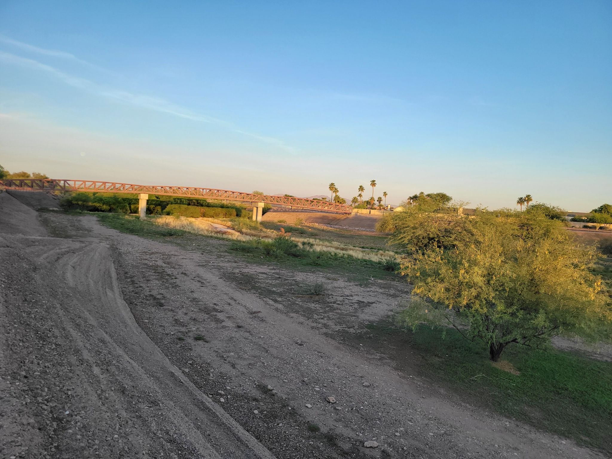 Dirt path leading towards a metal pedestrian bridge over dry ground at sunset. - Rio Vista Community Park – Arizona Canal Trail loop from Central Ave/Camelback