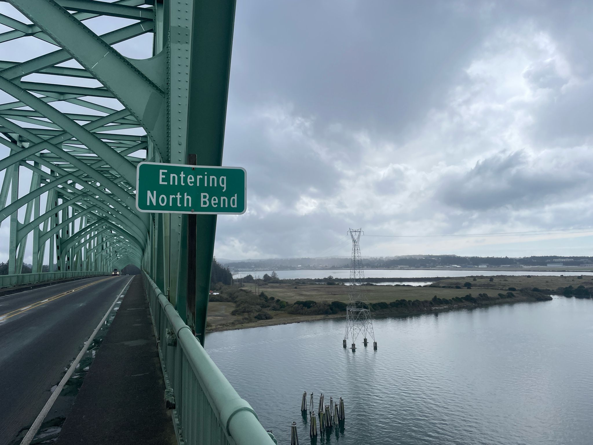 Green truss bridge over a wide river with distant land under a cloudy sky. - Oregon Coast Scenic Bikeway: Coos Bay Loop