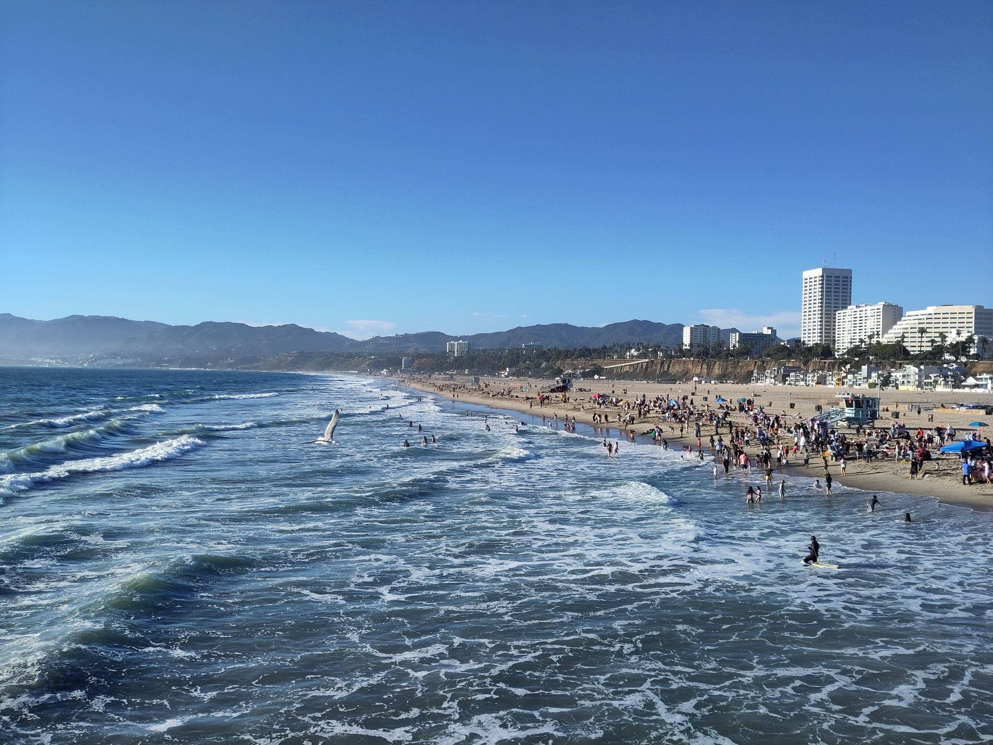 Crowded beach with ocean waves, city buildings, and mountains under a clear sky. - Santa Monica & Route 66 sign – Route 66: End of the Trail loop from Downtown Santa Monica