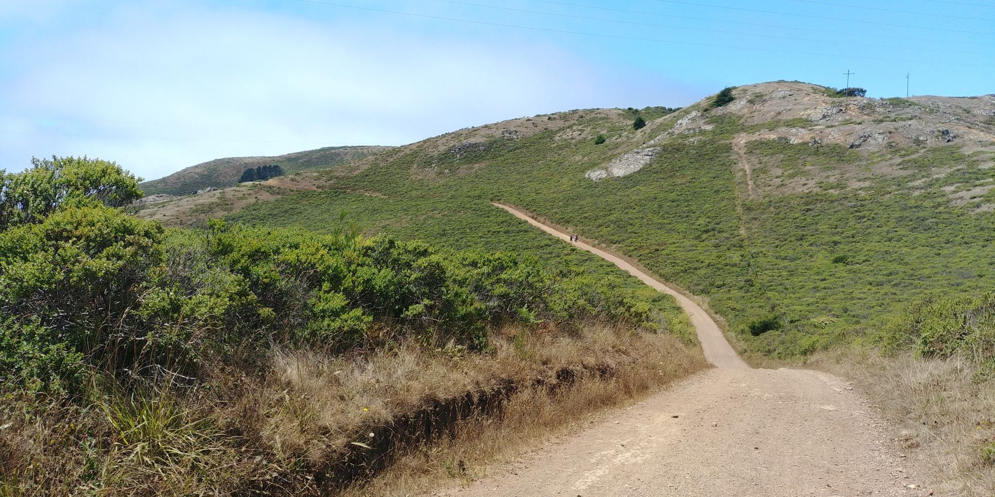 A winding dirt trail ascends a green, brush-covered hill with two distant hikers. - Alta, Bobcat and Miwok Trails Loop