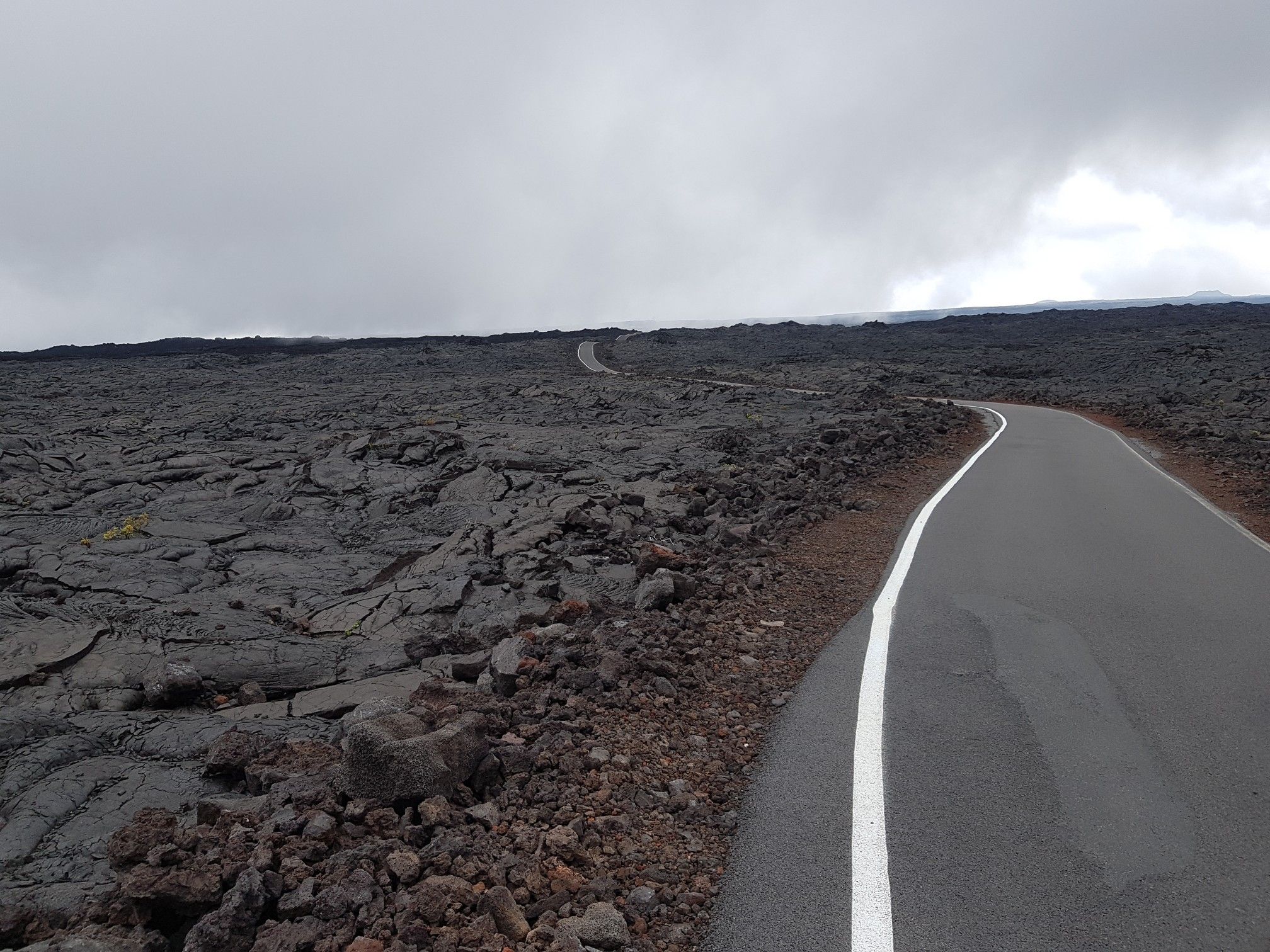 Paved road curves through a vast, dark volcanic field under an overcast sky. - Mauna Loa Observatory from Puʻu Huluhulu — Hawai‘i