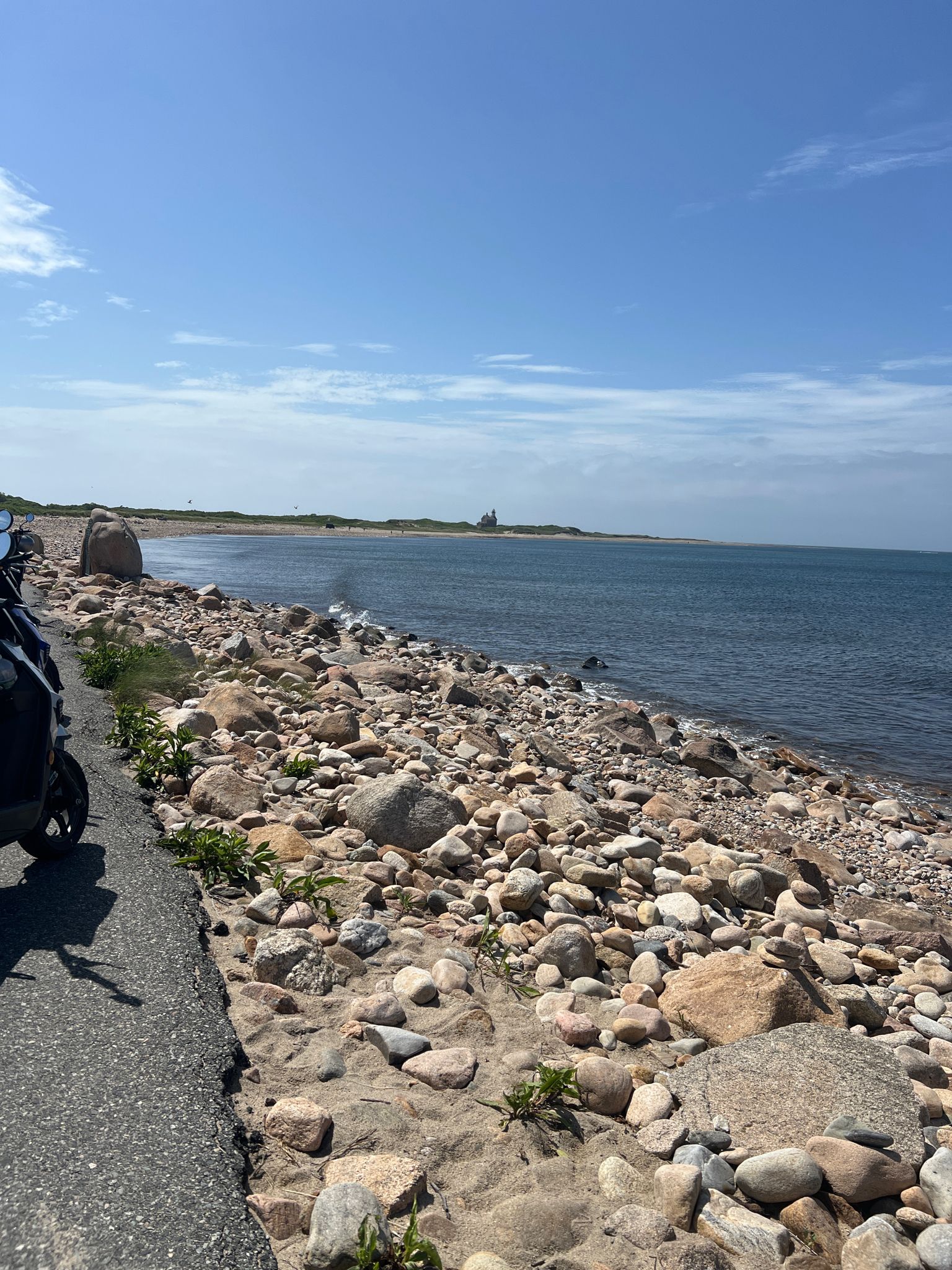 Rocky beach next to a paved path, leading to a distant coastline under blue sky. - North Lighthouse Trail