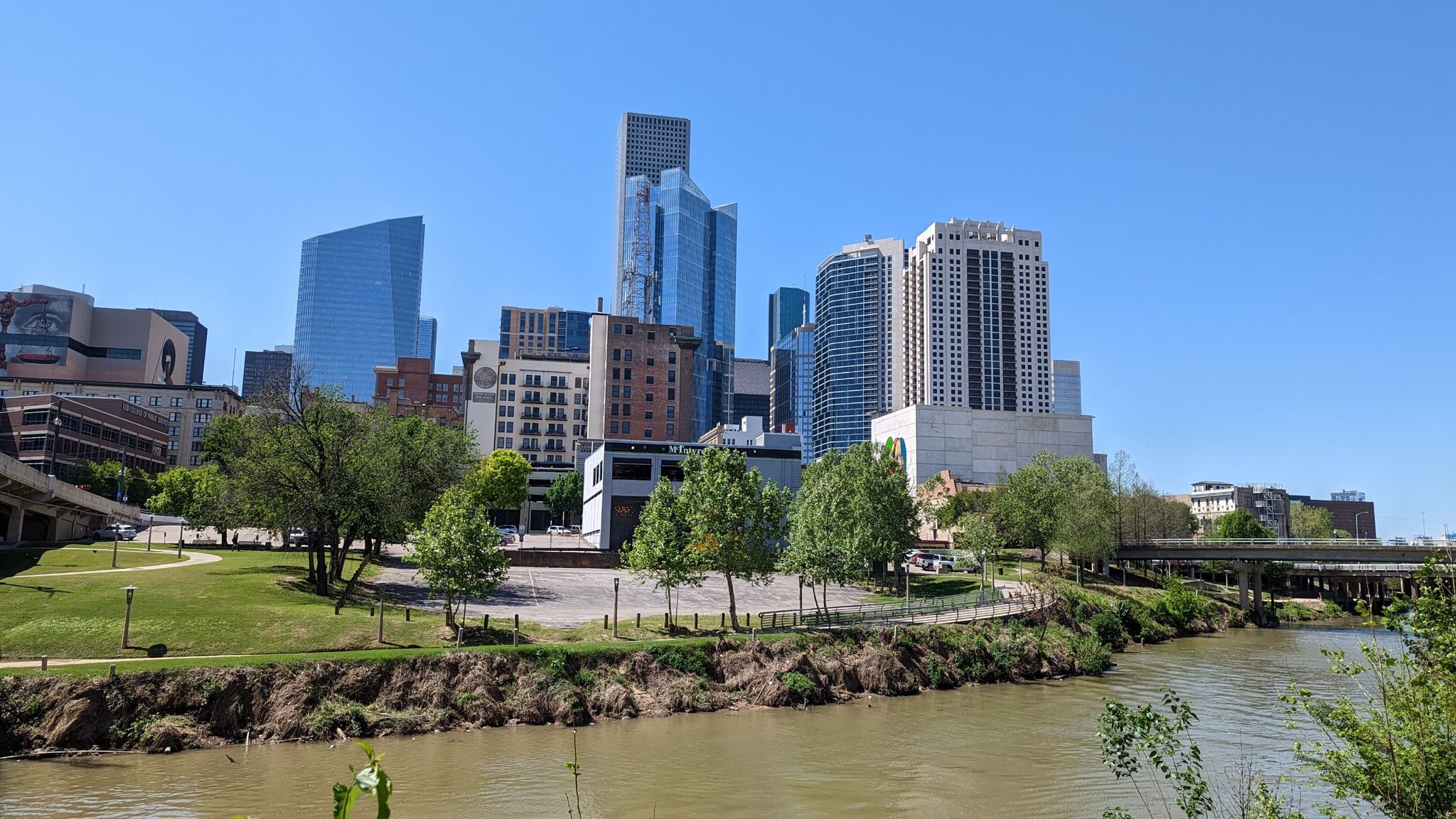Bright city skyline with tall buildings, green parkland, and a winding river. - The Cistern at Buffalo Bayou Park – Main Street Bridge loop from University of Saint Thomas