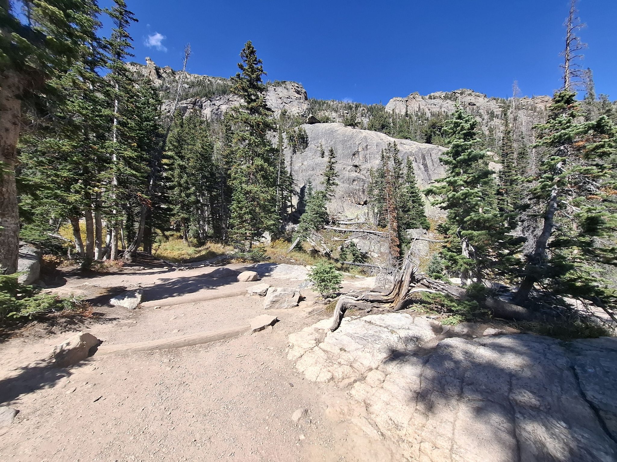 Dirt path through a sunny forest with large rock formations and mountains. - Emerald Lake Trail