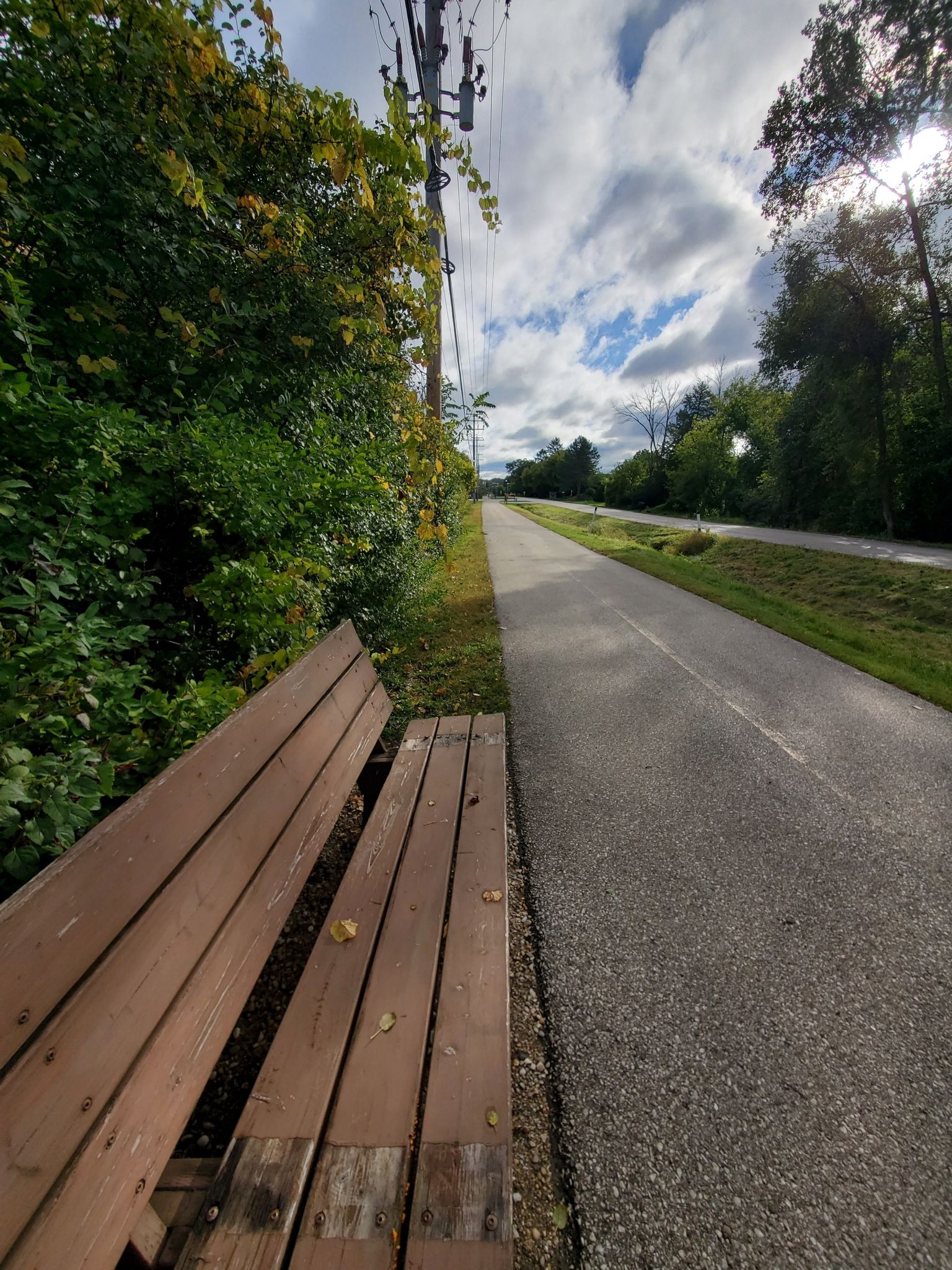 Empty wooden bench beside a paved path under a cloudy sky. - Nagawicka Lake loop from Village of Pewaukee