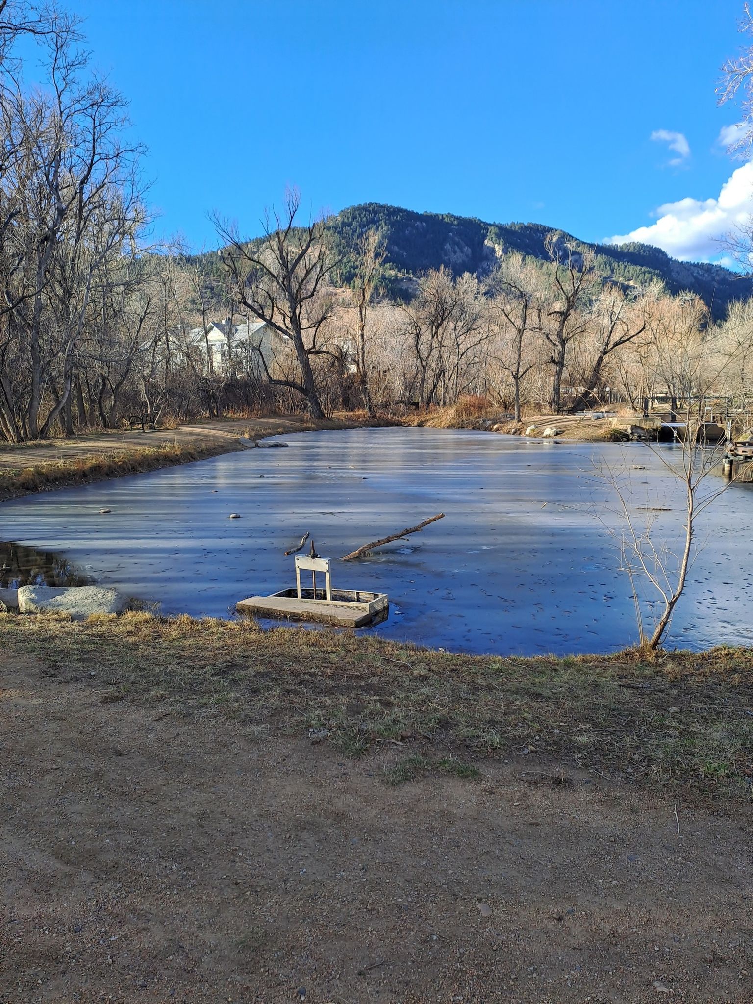 A partially frozen pond with bare trees and mountains under a blue sky. - Boulder Creek Path loop from Boulder
