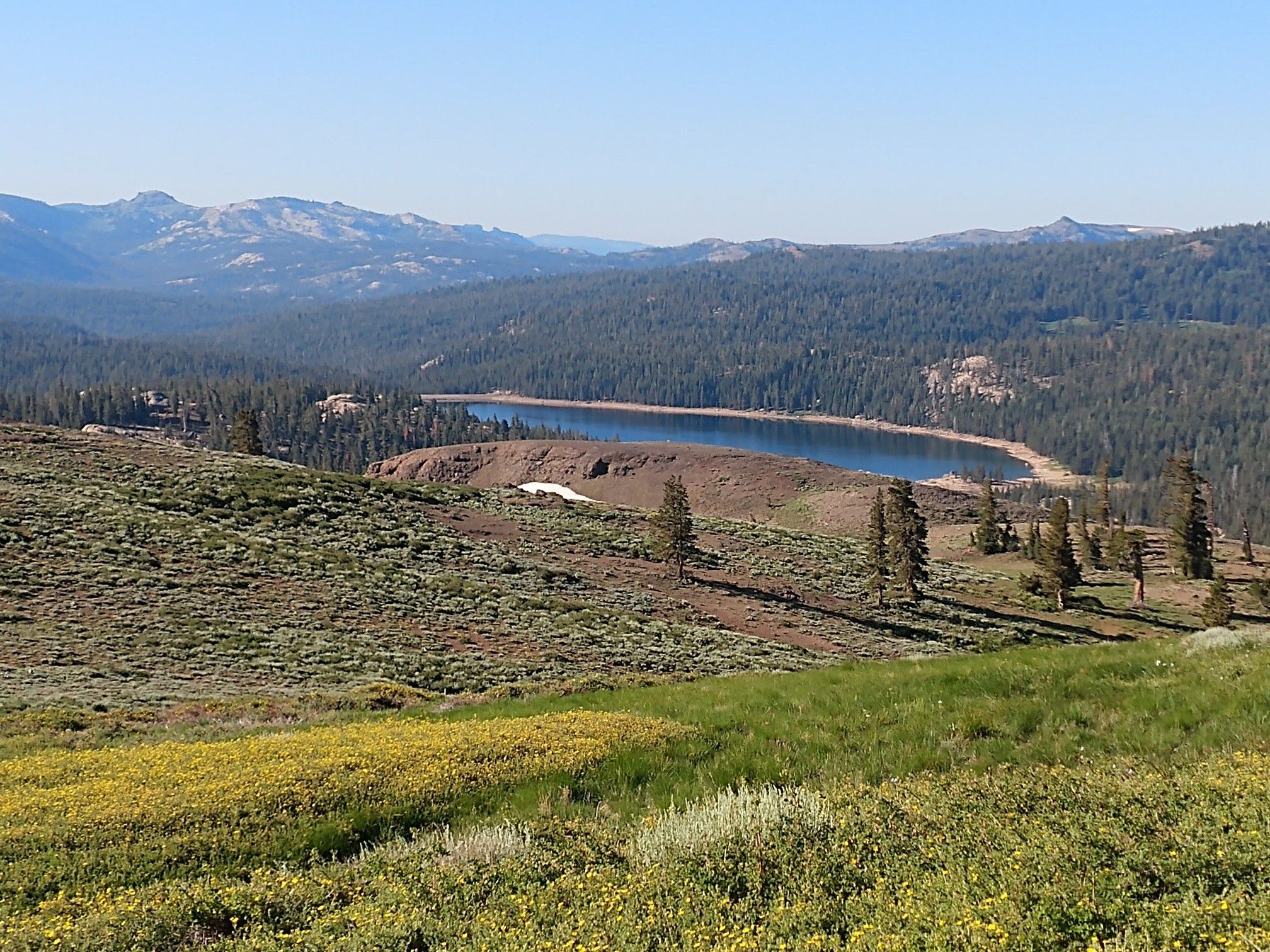 A scenic lake surrounded by forested hills and distant mountains under clear skies. - The Nipple Trail