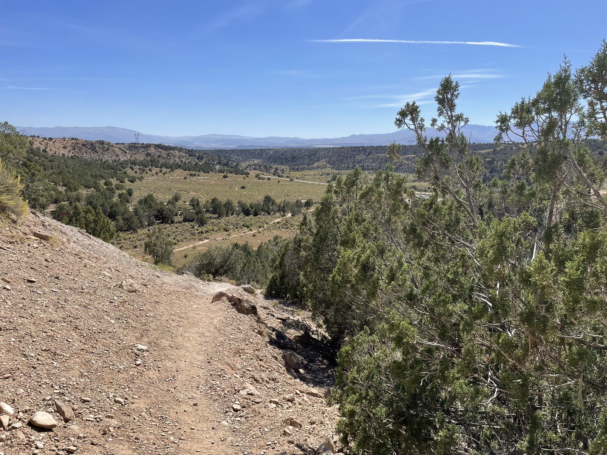 A dirt path winds down an arid slope overlooking a green valley. - Rifle Arch Trail