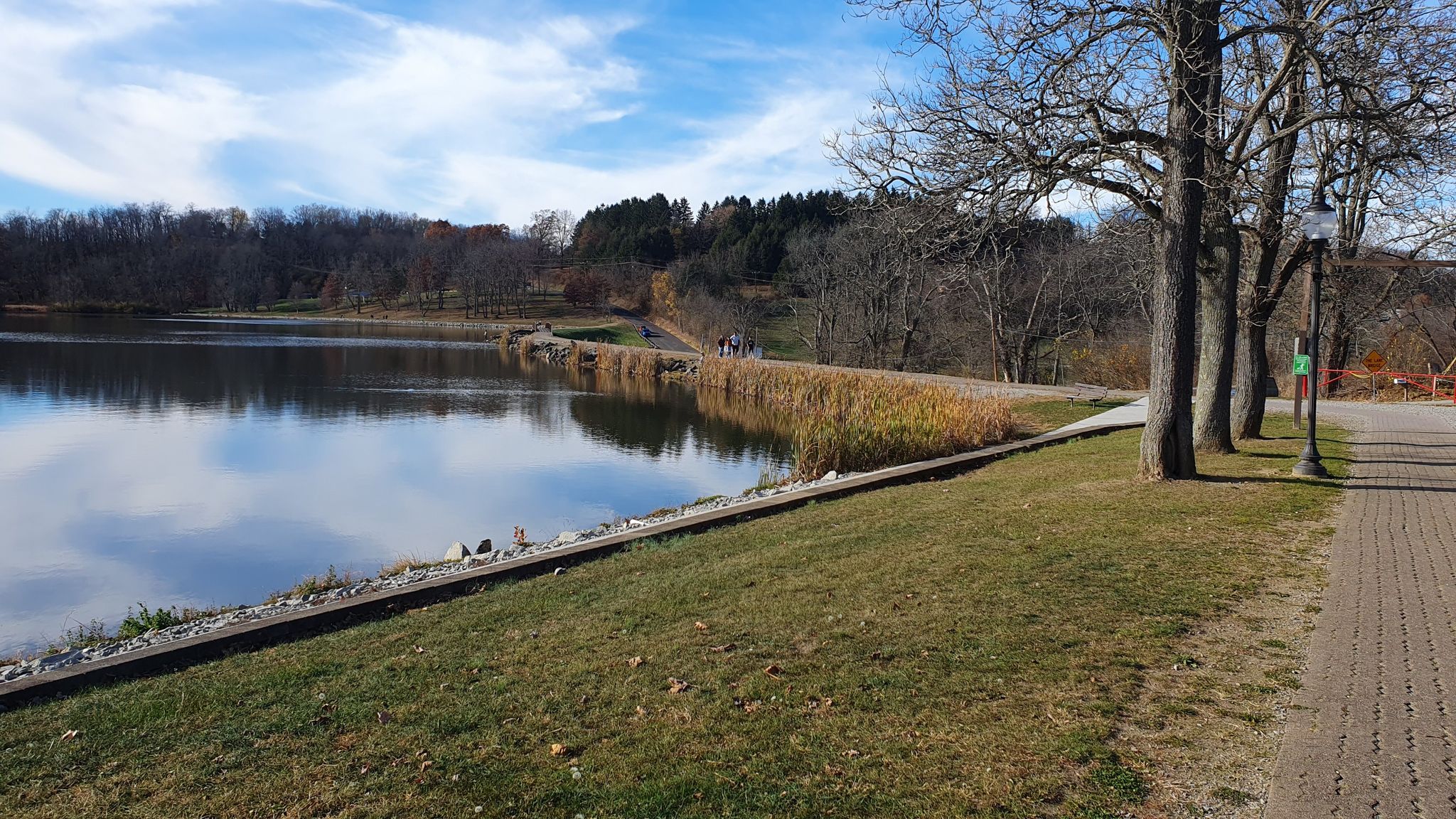 Calm lake reflecting blue sky, with a paved path and distant walkers. - Twin Lakes Park Loop