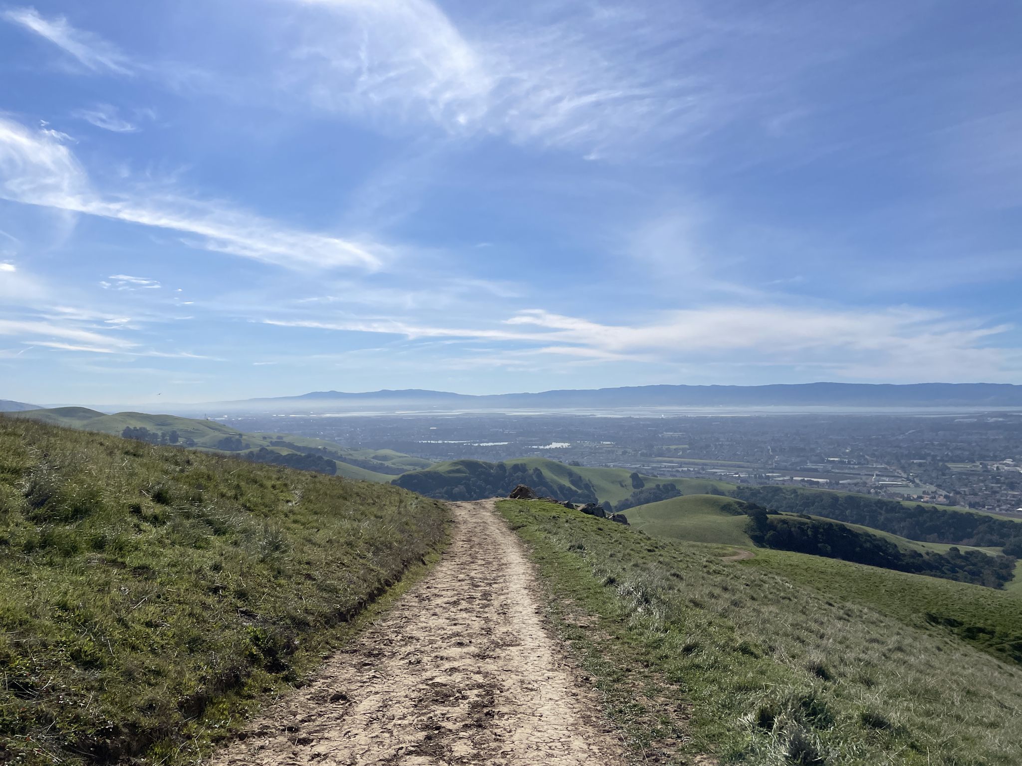 Dirt path through green hills with a distant city under a bright blue sky. - High Ridge and Gossip Rock Loop