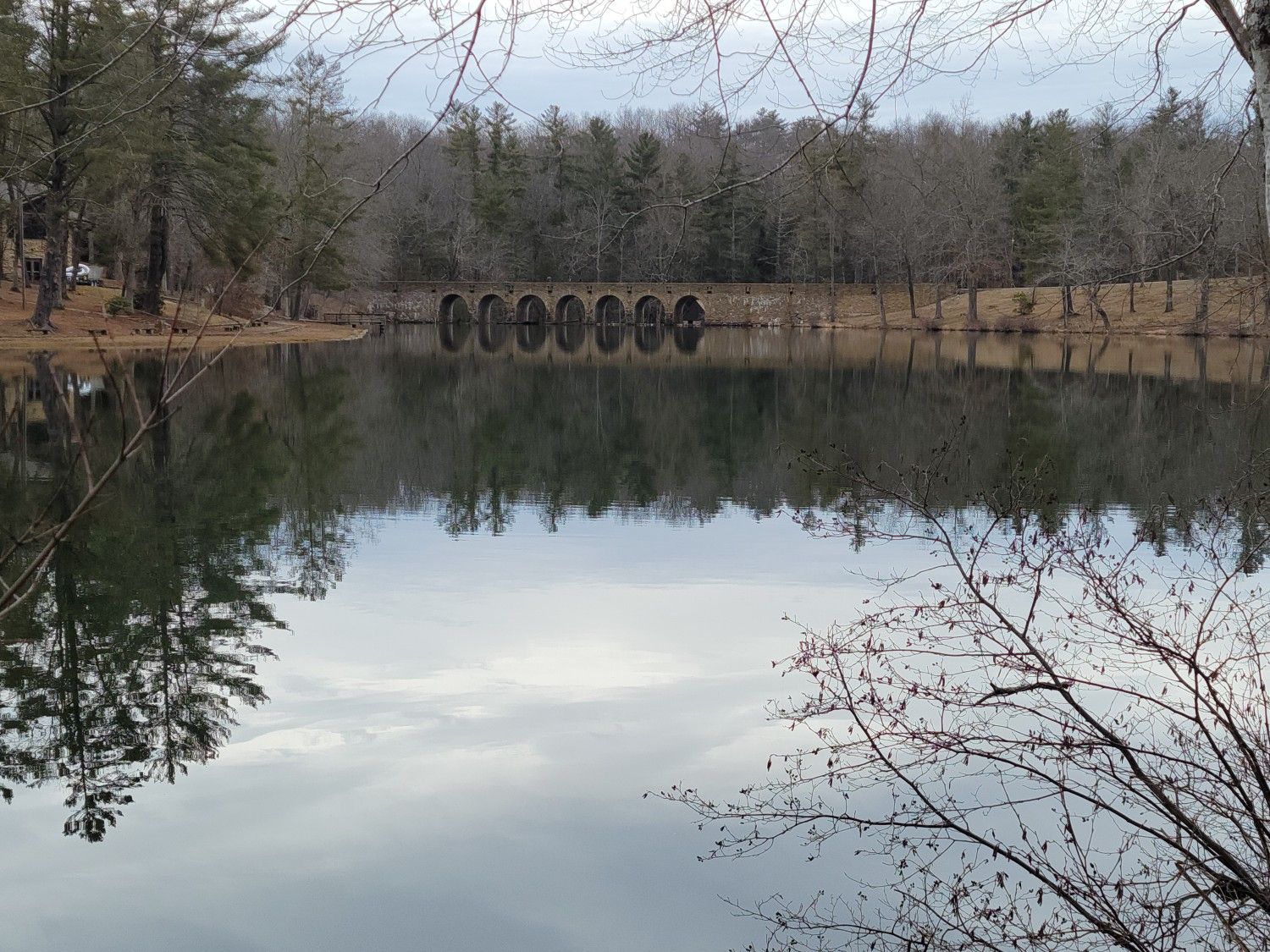 Calm lake reflecting bare trees and an arched stone bridge under a cloudy sky. - Byrd Lake loop from Cumberland Mountain State Park