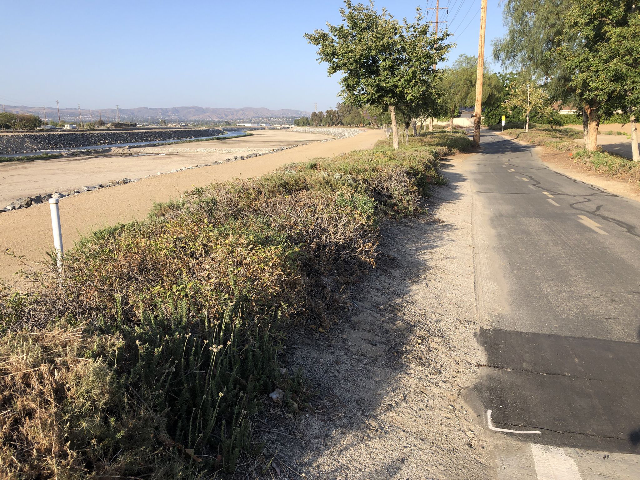 Paved path next to dense green and brown bushes, with a dry riverbed. - View of Sierra Peak – Santa Ana River Trail Bridge loop from Anaheim Canyon