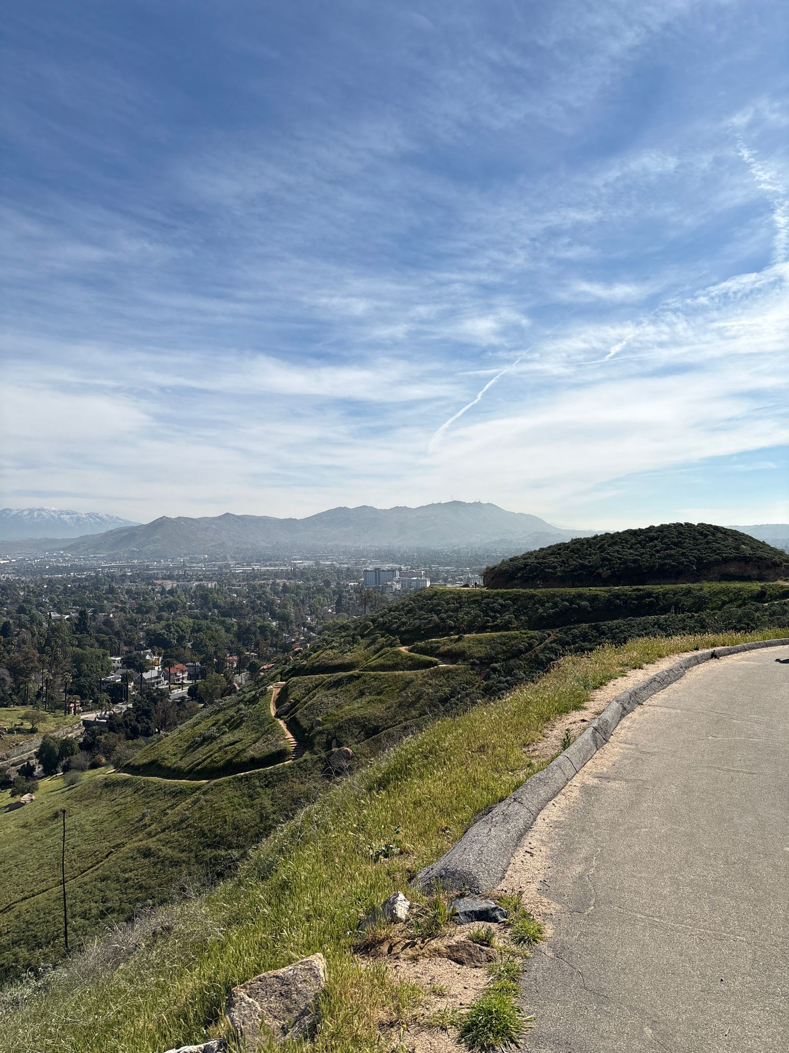 A winding road on a green hillside overlooks a vast valley and distant mountains. - Bark Park – Mount Rubidoux loop from Riverside