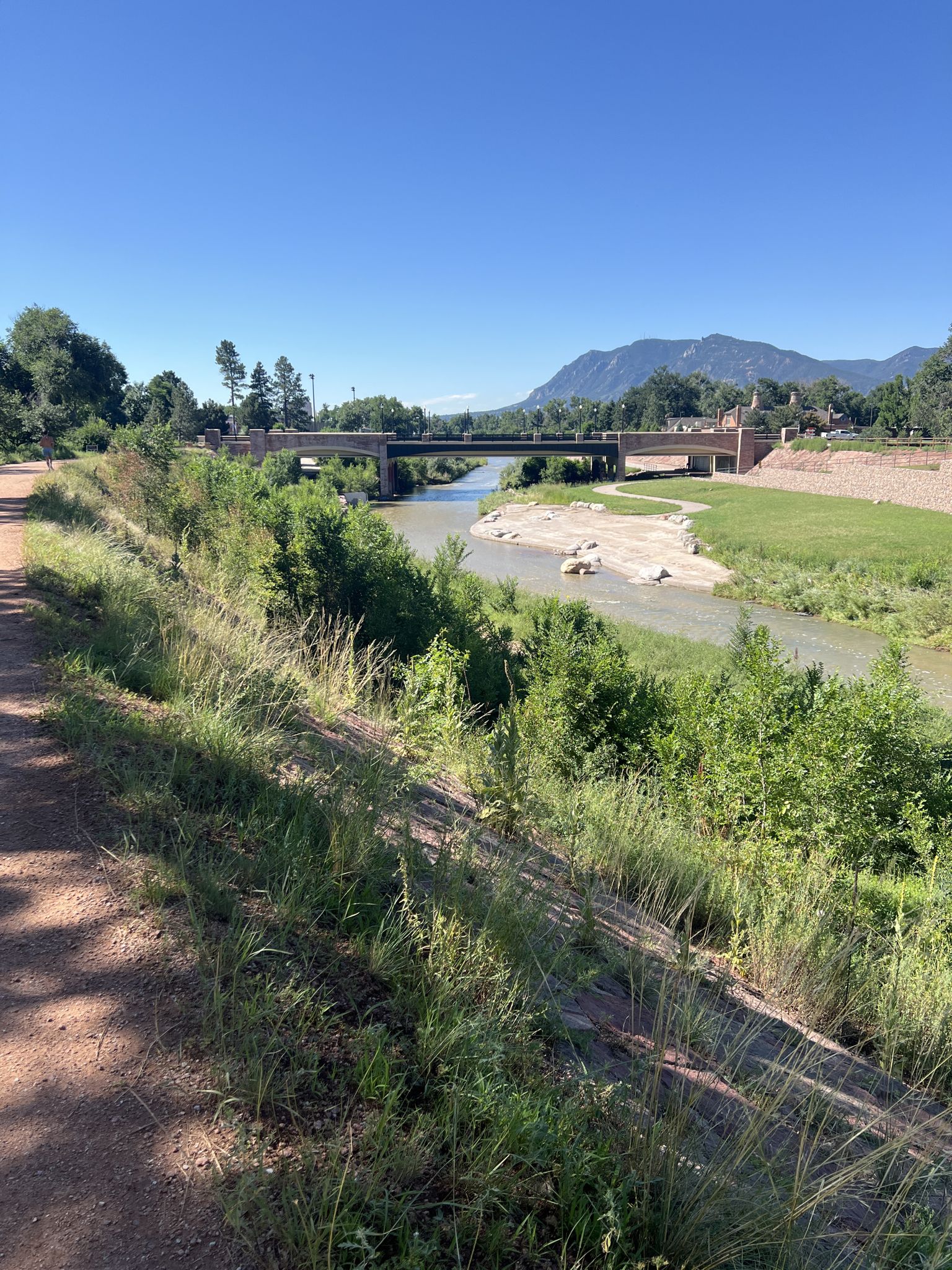 Dirt path overlooking a river with a bridge, green trees, and distant mountains. - Fountain Creek Regional Trail – Uintah street bridge loop from KCSF-AM (Colorado Springs)
