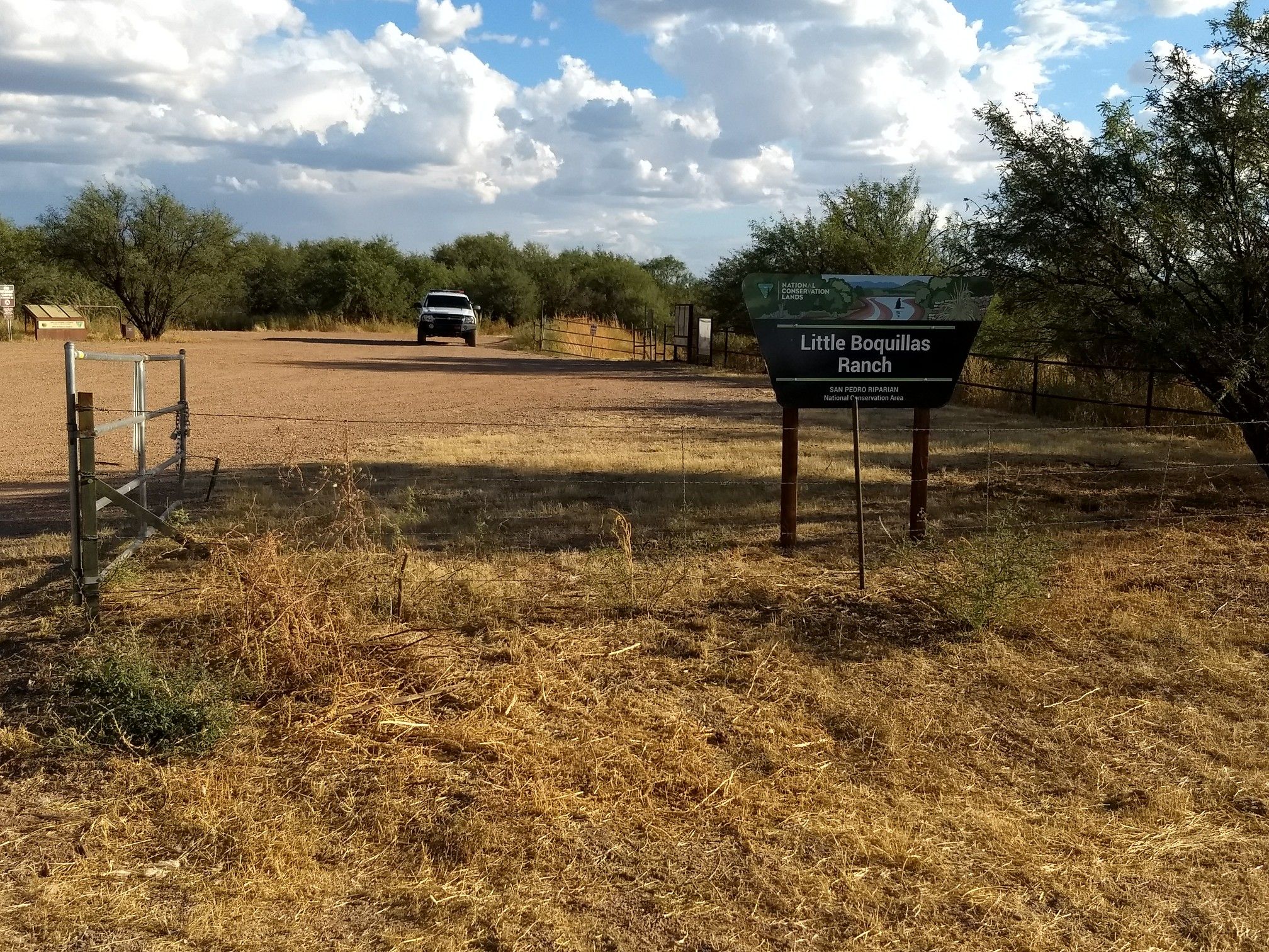 Dirt parking area with a sign, distant vehicle, and sparse trees under cloudy sky. - Fairbank Loop via San Pedro Trail