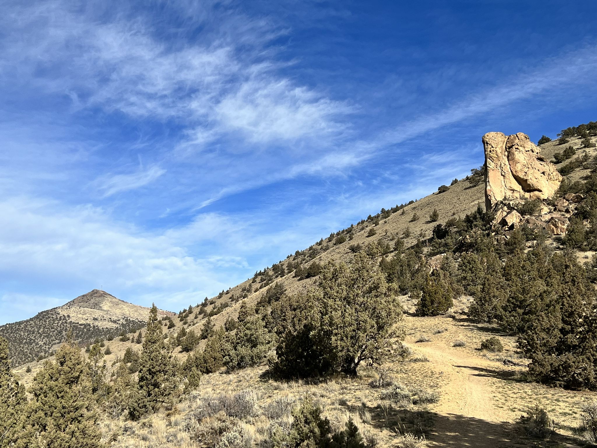Dirt path winds up a dry hillside past a large rock under a blue sky. - Gray Butte Trail