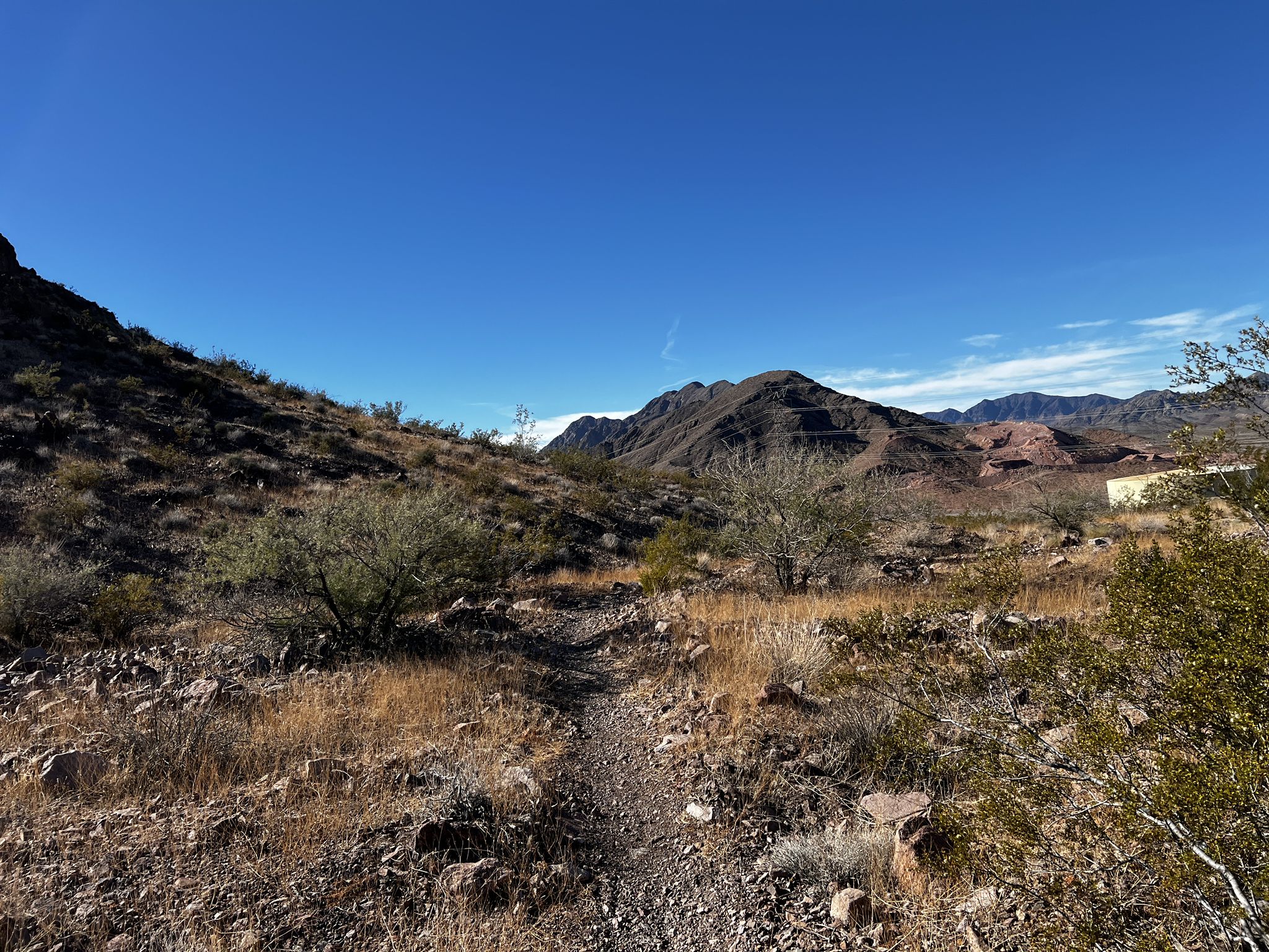 A narrow dirt path winding through arid scrubland with rocky hills under a blue sky. - Old Vegas to Railroad Pass Trail