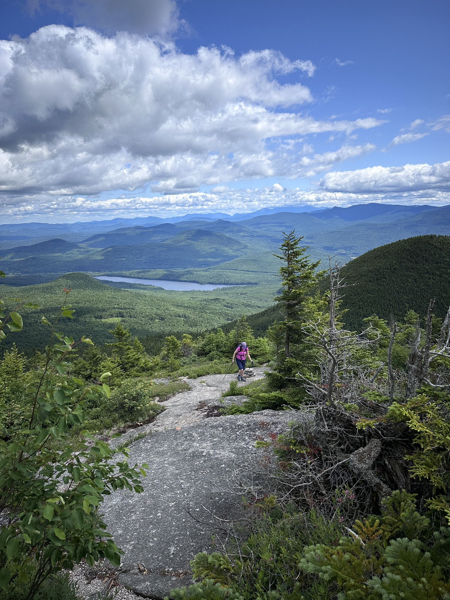 Hiker on a rocky path overlooking a vast mountain landscape with a lake. - Percy Peaks Trail