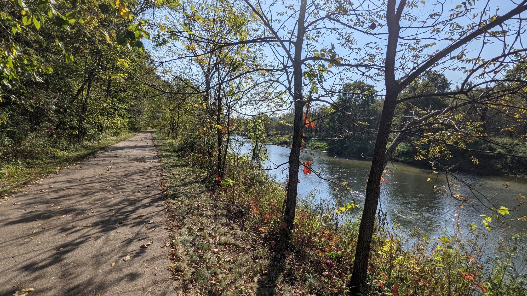 Paved path beside a sunlit river, surrounded by autumn trees. - Peterson sign with a gnome – Barn and Pump House loop from Lanesboro