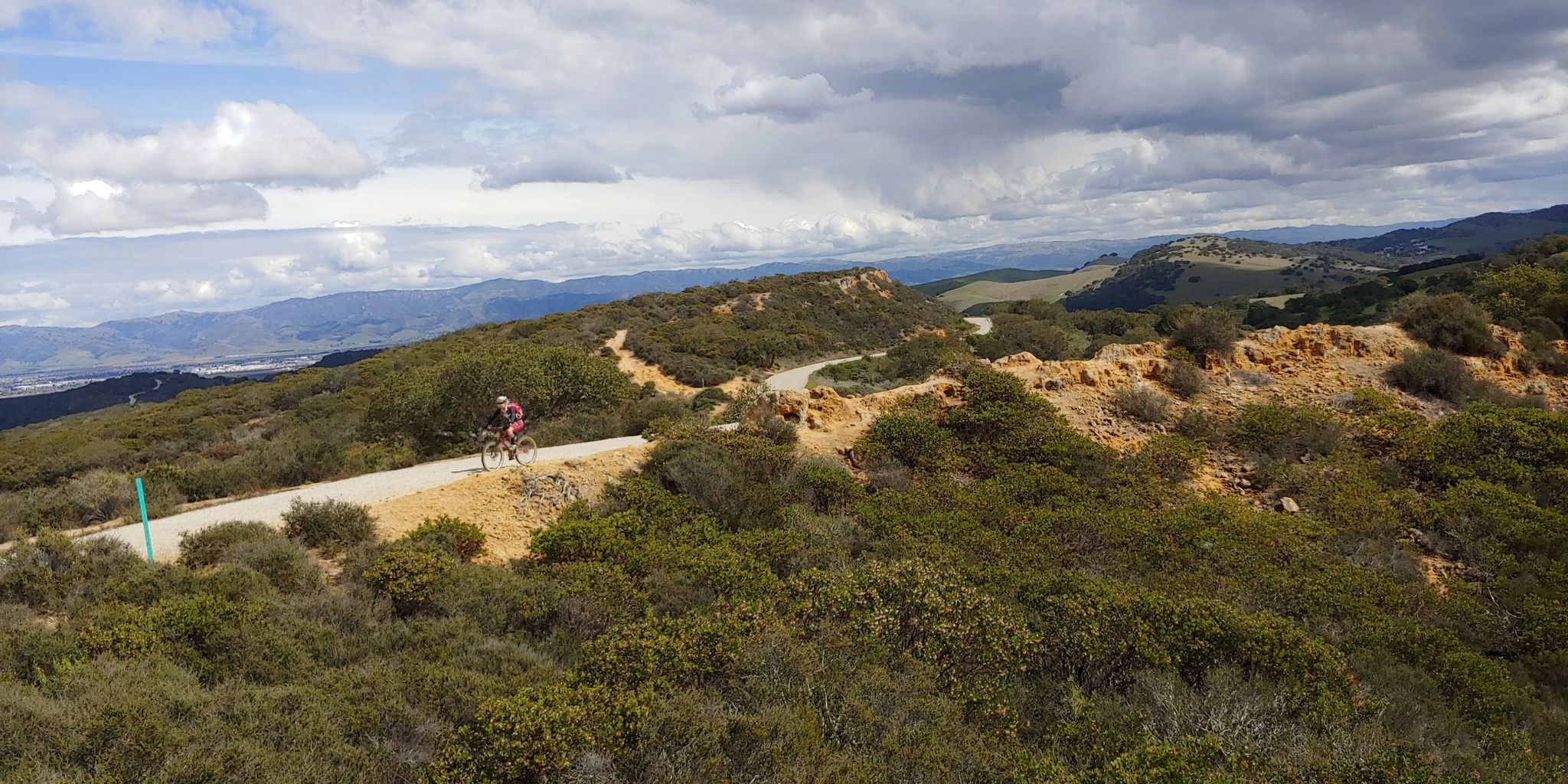 A cyclist rides a winding dirt road through brushy hills under a cloudy sky. - Trail 41 and Trail 36 Loop