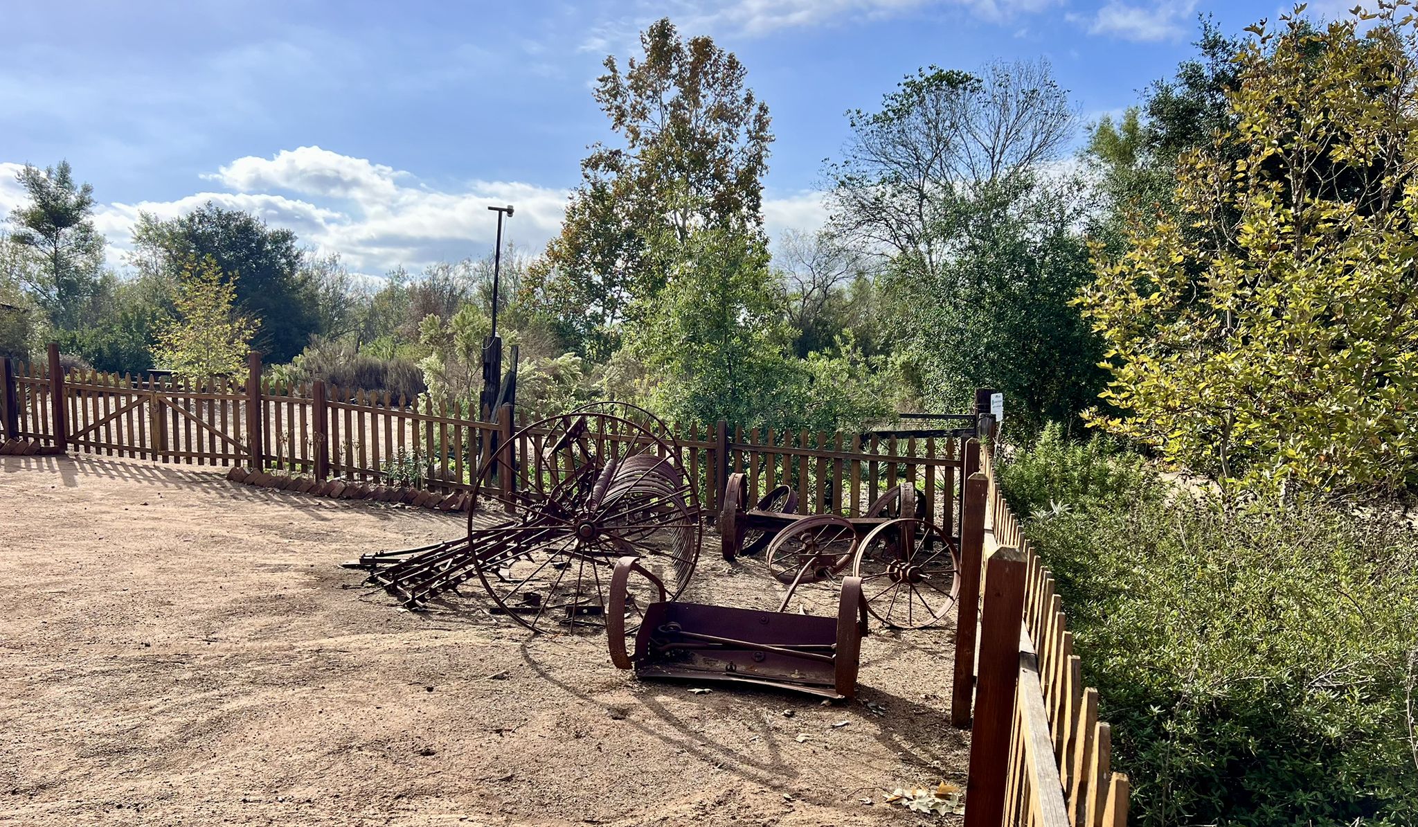 Old rusty farm equipment sits in a fenced area with trees and blue sky. - Sikes Adobe Historic Farmstead – View of I-15 loop from Westfield North County