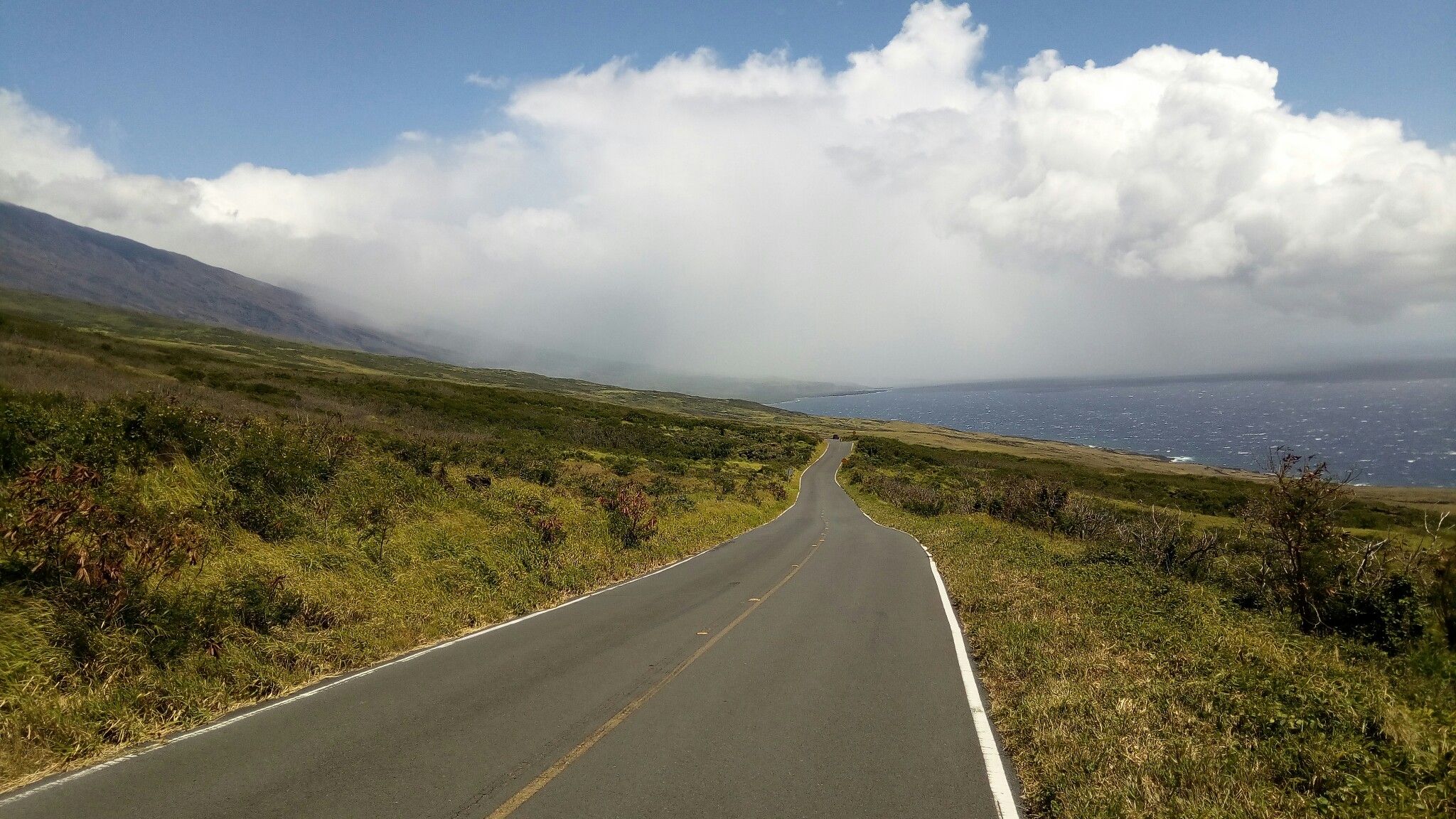 Paved road winding through green hills towards a cloudy ocean horizon. - Kepuni Rocks Viewpoint loop from Keokea