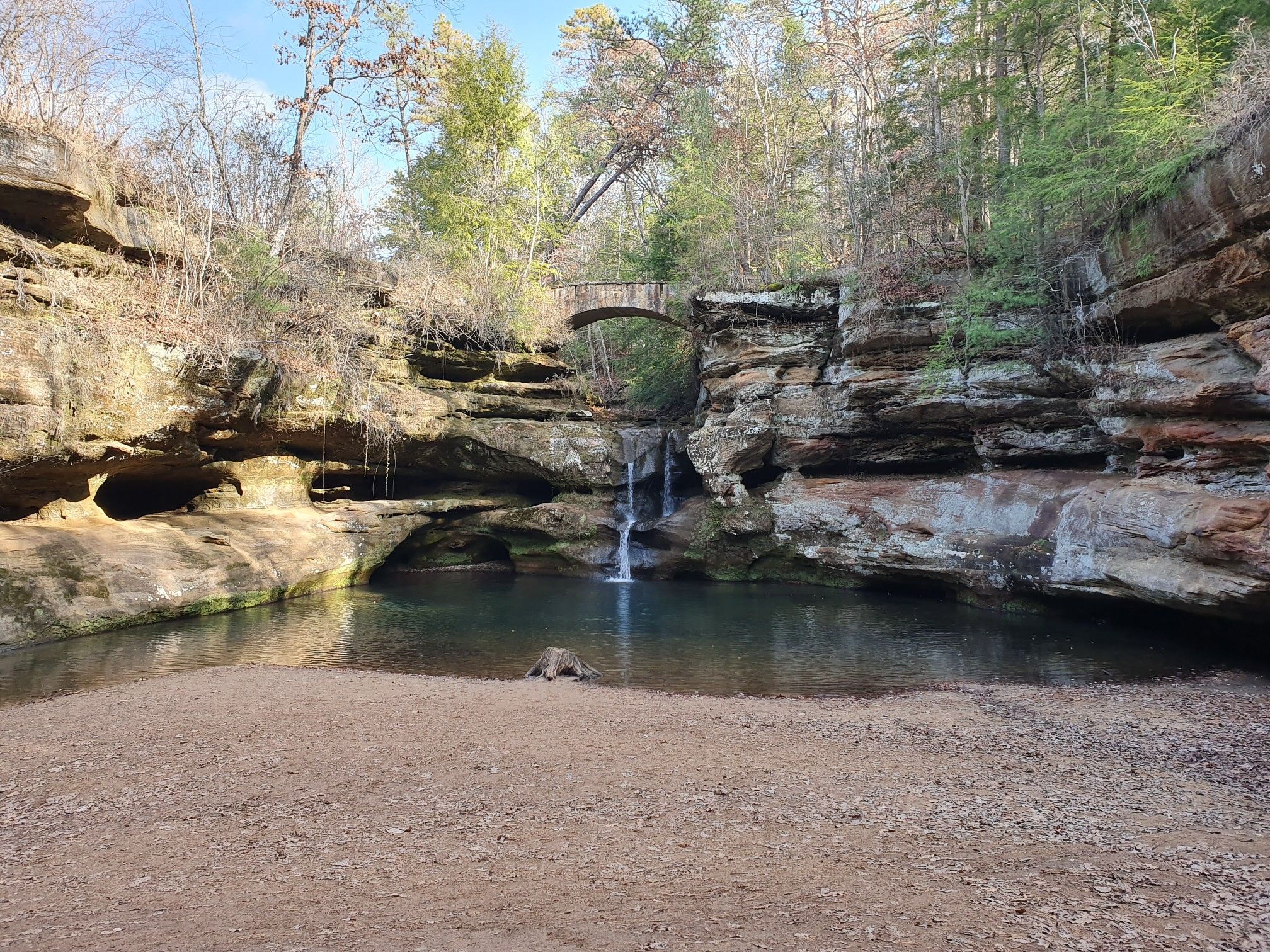 Waterfall flowing into a clear pool, with a stone arch bridge overhead. - Old Man's Cave to Whispering Cave Loop